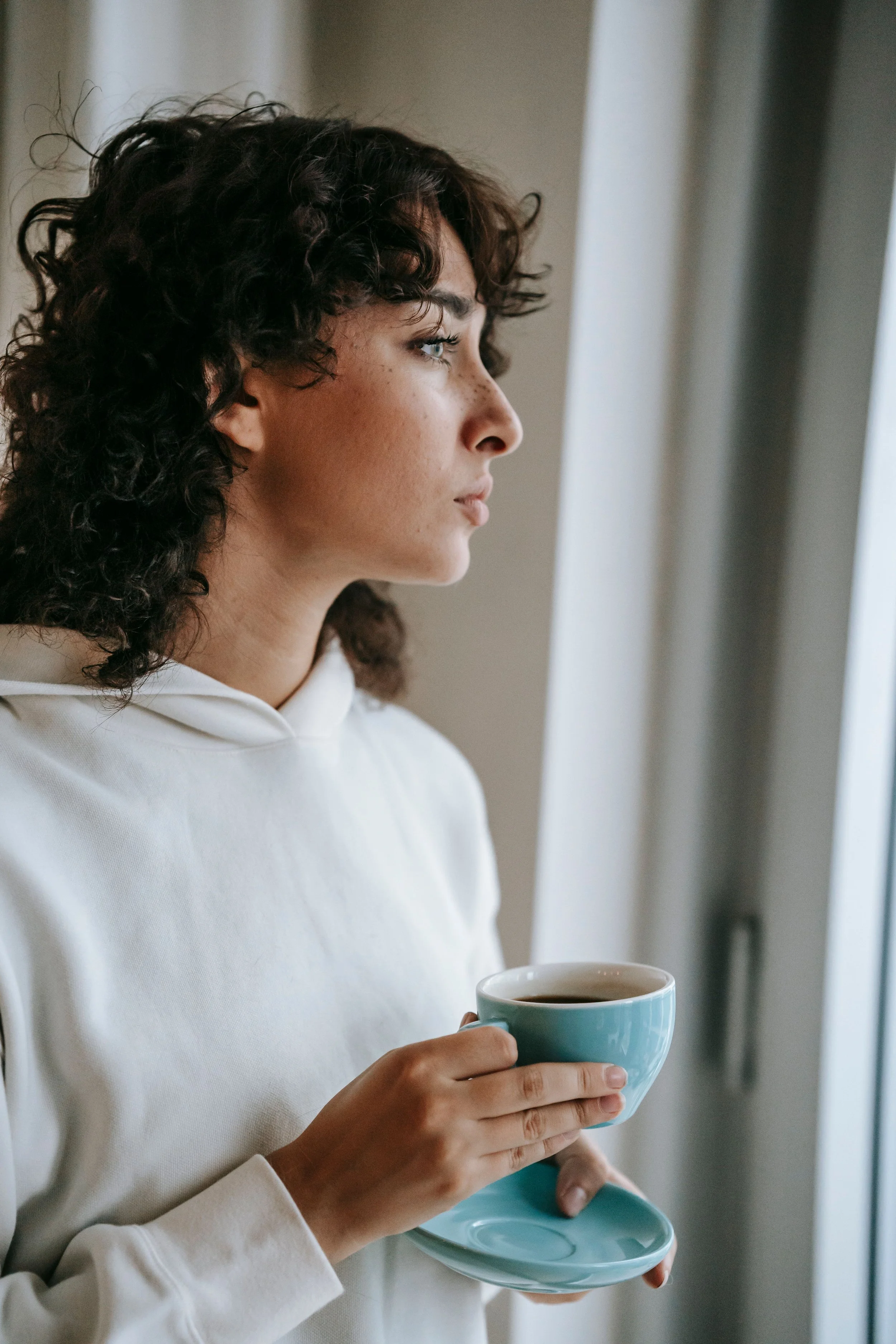 A woman with curly hair holding a cup of coffee or tea and a saucer, standing near a window.