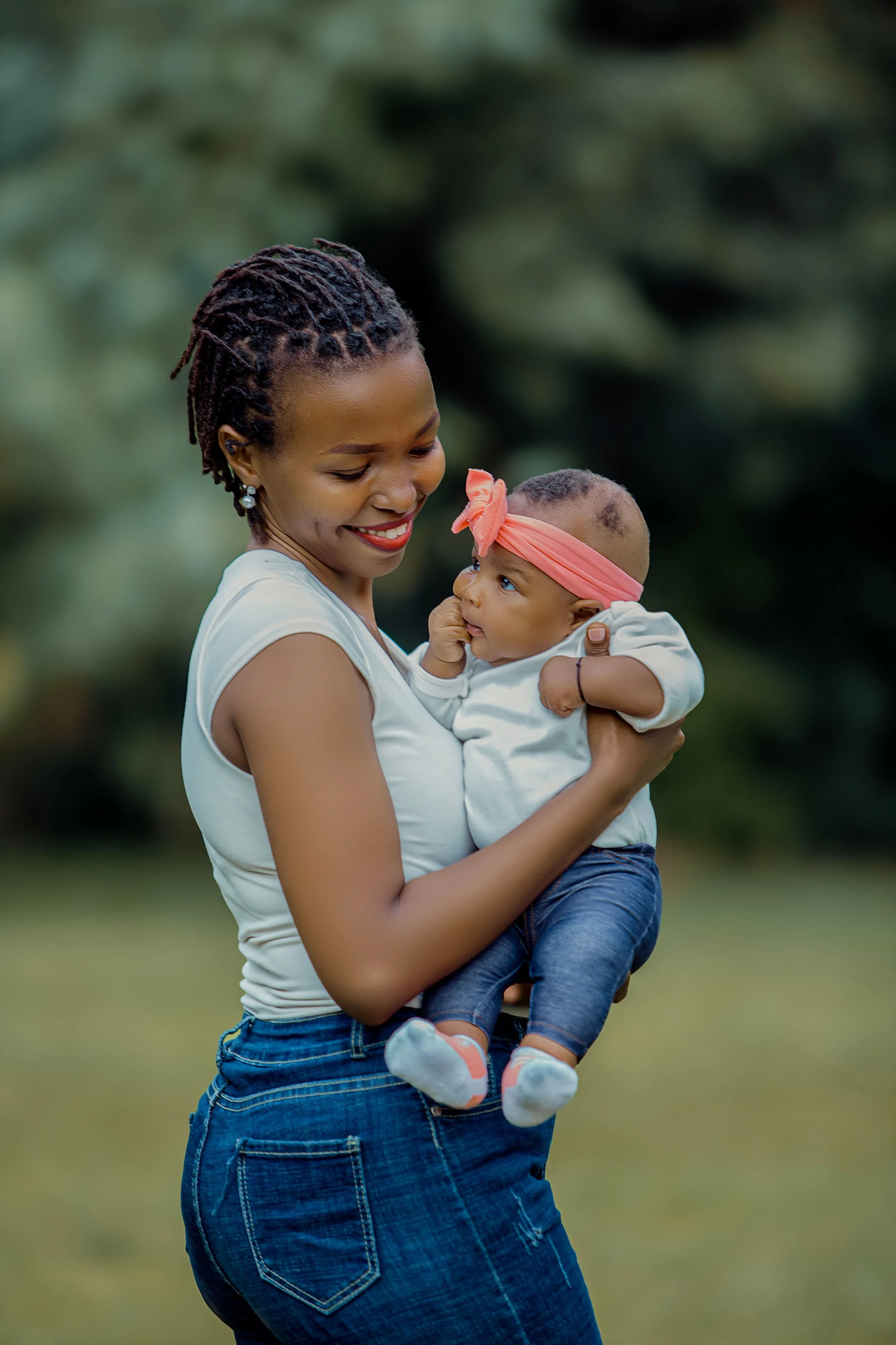 A woman holding a baby outdoors in a park or garden with a blurred green background. The woman is smiling and looking at the baby, who is wearing a pink headband and gazing at her.