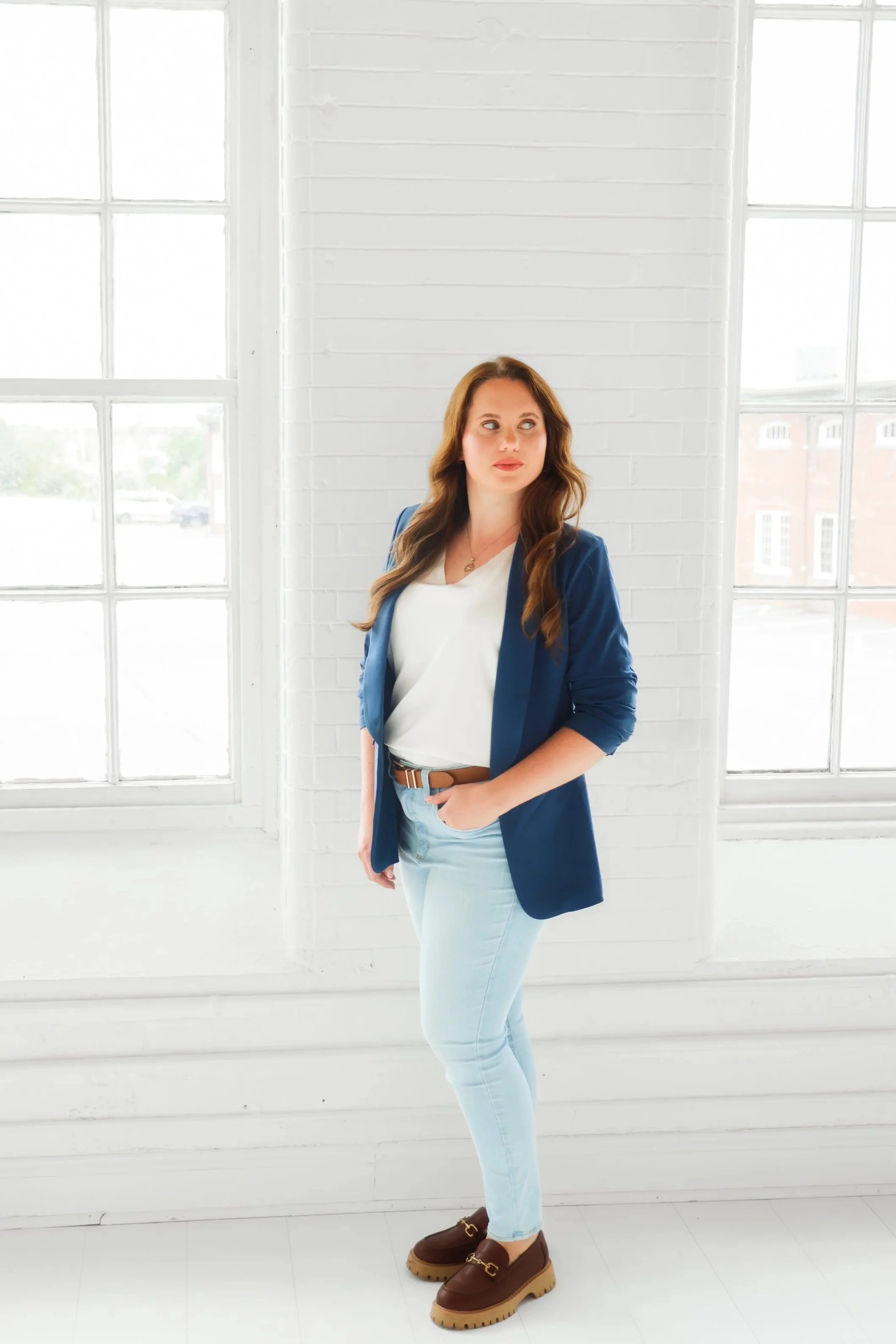A woman with long brown hair wearing a white top, denim jeans, a blue blazer, and brown platform shoes standing indoors with white brick walls and large windows behind her.