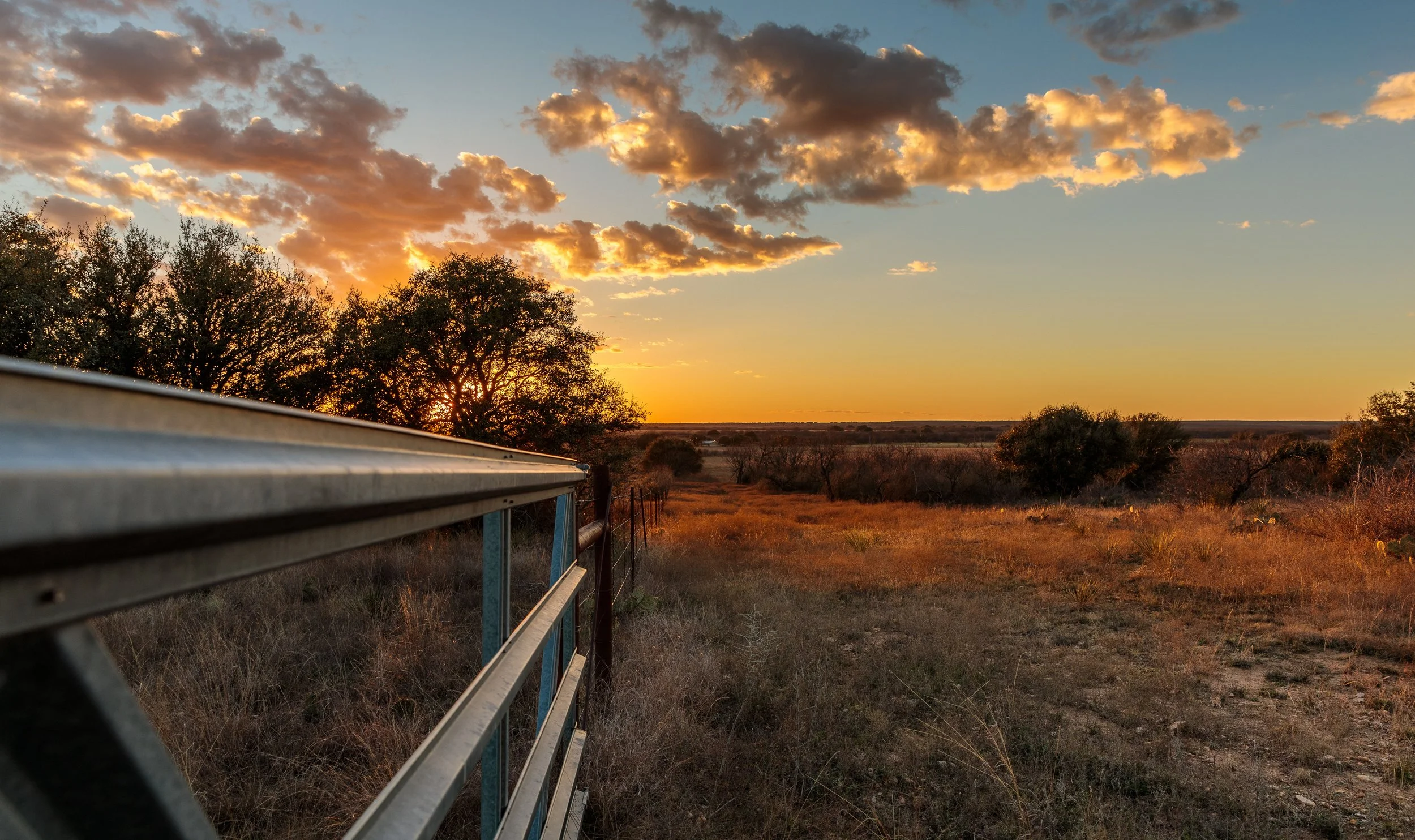 Sunset over a rural landscape with trees, open fields, and a metal railing in the foreground.