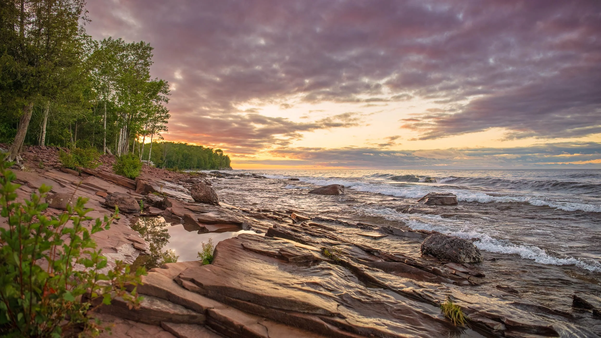 A rocky lakeshore at sunset with waves gently crashing on the rocks, a reflection pool in the foreground, and trees lining the shore on the left. The sky is partly cloudy with warm colors from the setting sun.