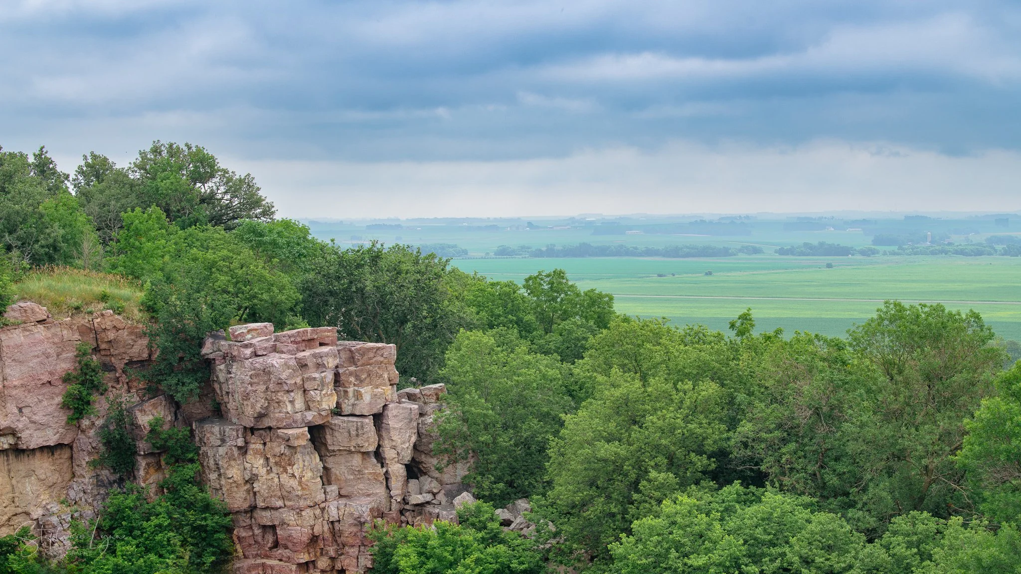 Scenic landscape of a forested area with rocky cliffs in the foreground and open green fields and distant farmland under a cloudy sky in the background.