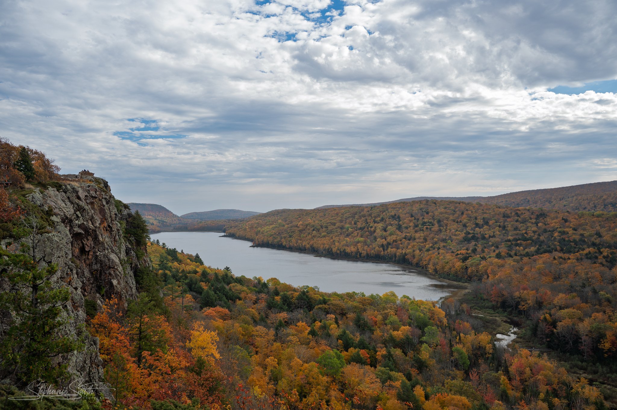 A river flowing through a valley surrounded by autumn-colored trees, with hills and cliffs on the sides, under a cloudy sky.