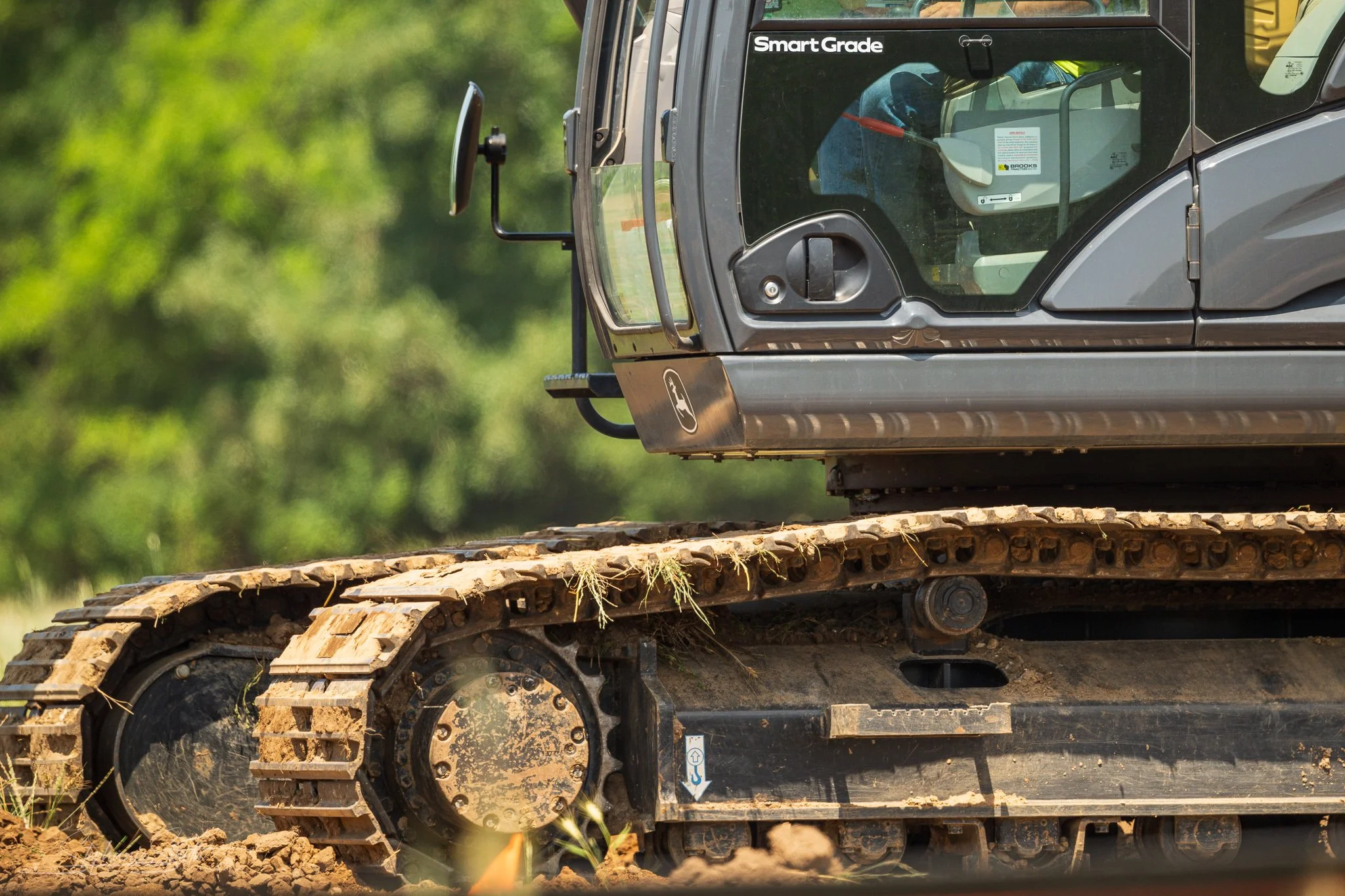 Close-up of a gray construction vehicle, specifically an excavator or bulldozer, with tracked wheels and a glass cabin, on a dirt surface with green blurred trees in the background.