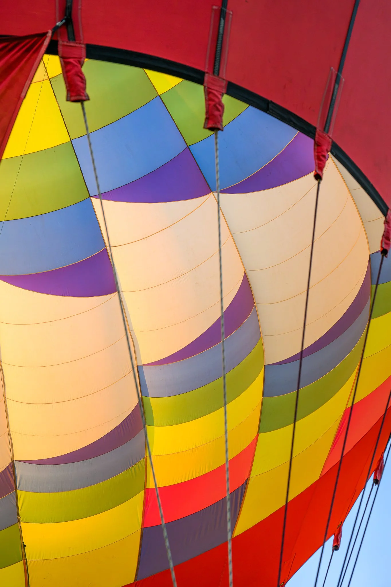 Close-up view of a multicolored hot air balloon fabric with red, yellow, purple, green, and beige sections, supported by thin ropes and metallic structural elements.