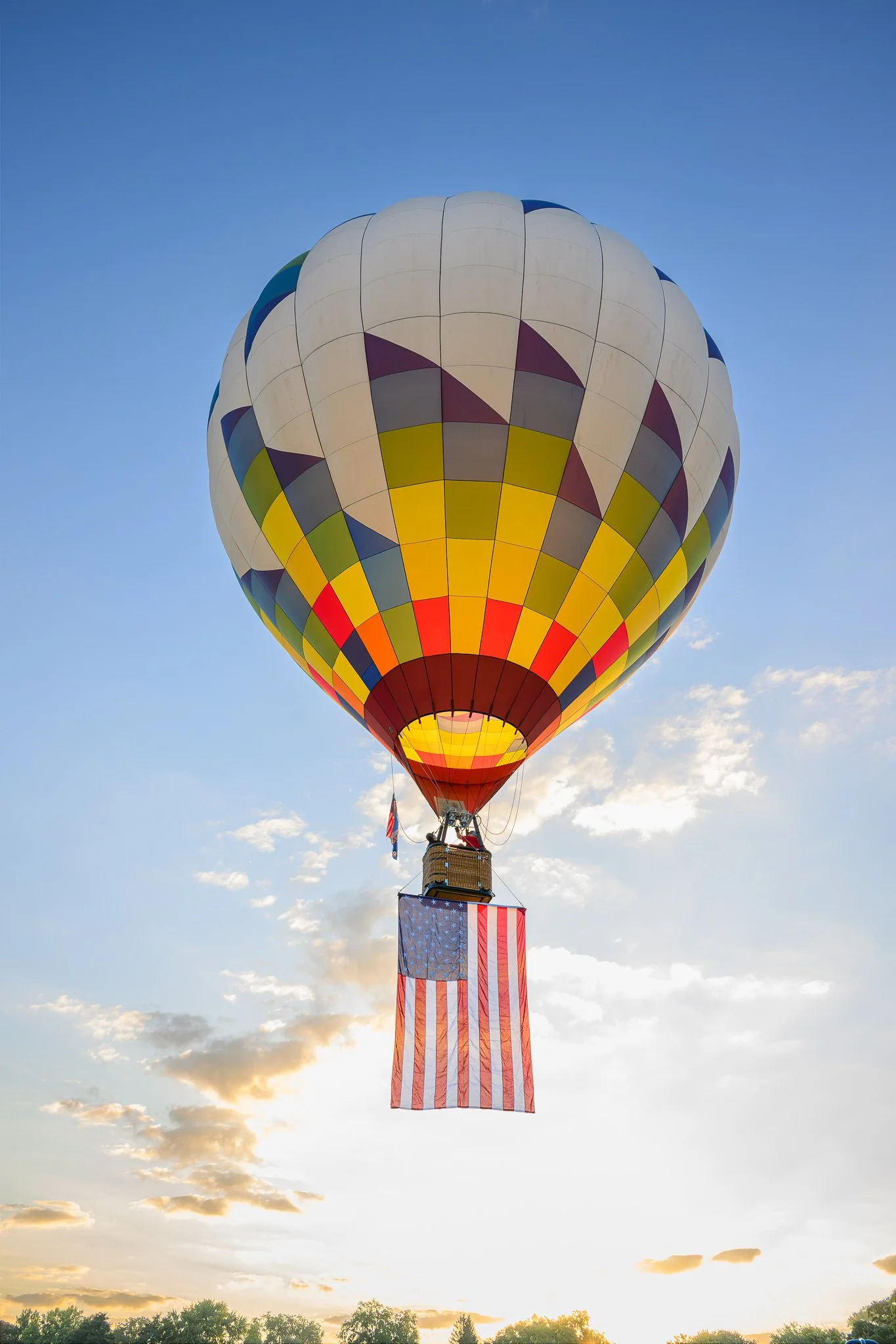 A hot air balloon decorated with an American flag, flying in the sky during sunset, with trees at the bottom.