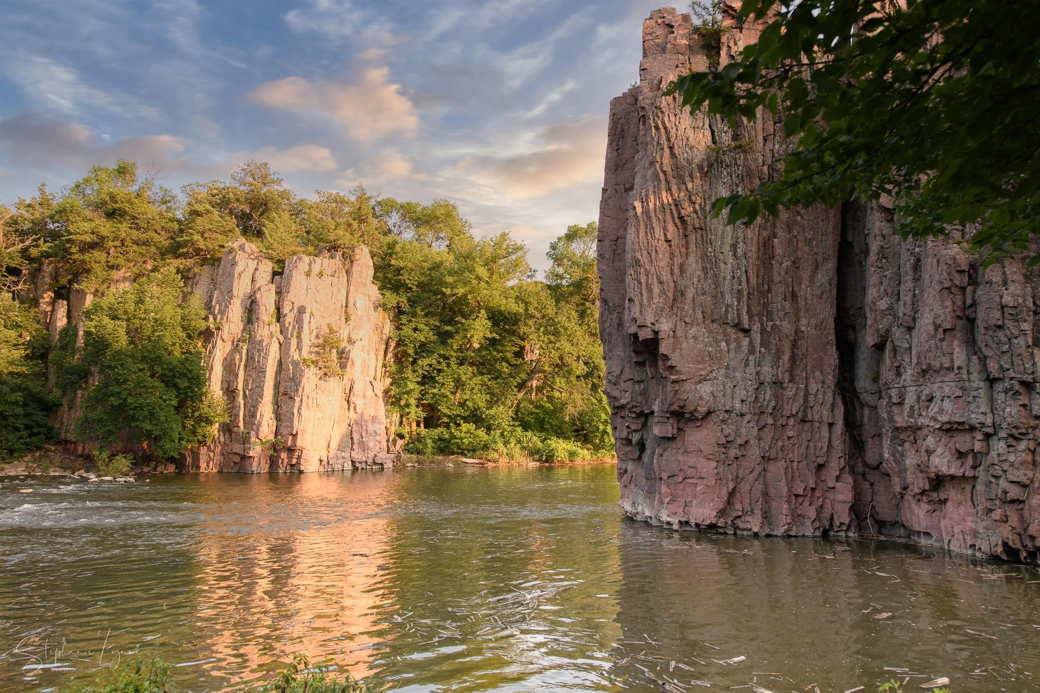 A river flowing through a canyon with tall, rugged rock formations and lush green trees. The sky is partly cloudy with a hint of sunlight.