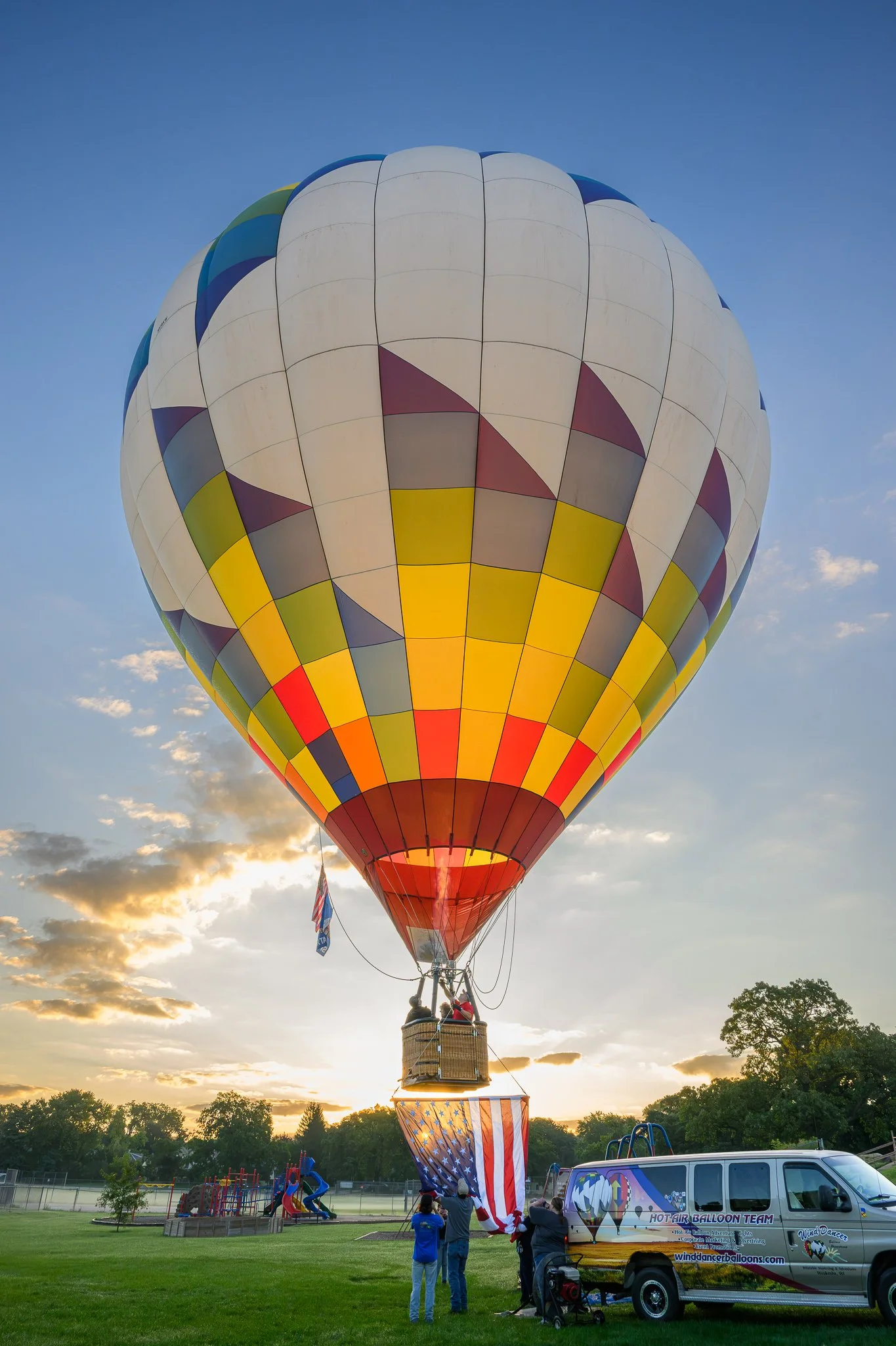 A hot air balloon with a colorful pattern of yellow, gray, purple, blue, and white panels preparing for flight in a park at sunset. Several people are standing nearby, and a van with a logo of a hot air balloon team is parked next to the balloon.