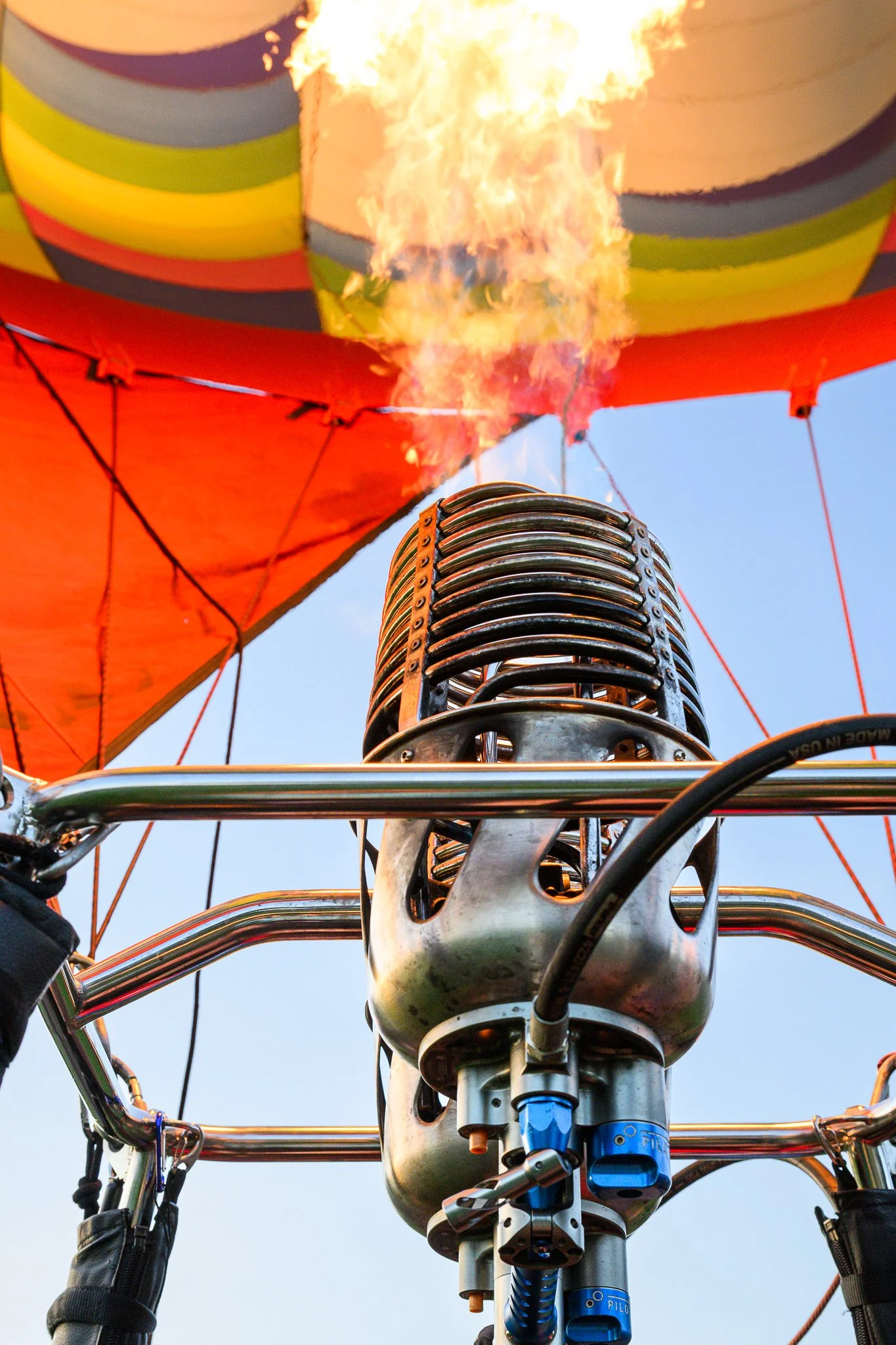 View from below of a hot air balloon with flames coming from the burner, which is mounted on the basket framework. The balloon has rainbow-colored stripes, and the sky is clear and blue.