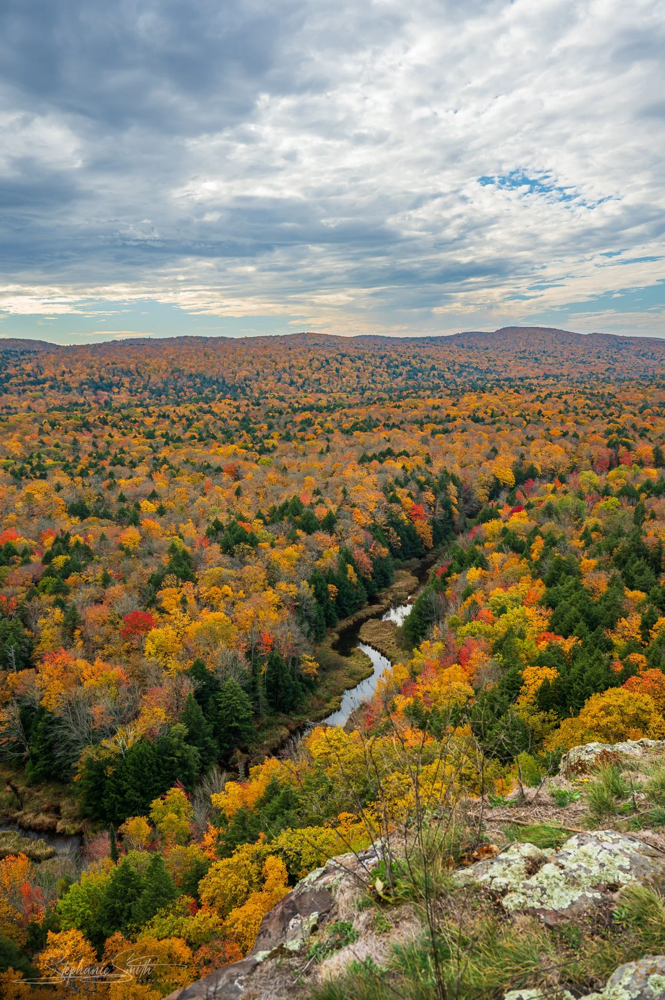 A scenic view of a forested mountain landscape in autumn, with colorful fall foliage and a winding river in the valley below, under a partly cloudy sky.