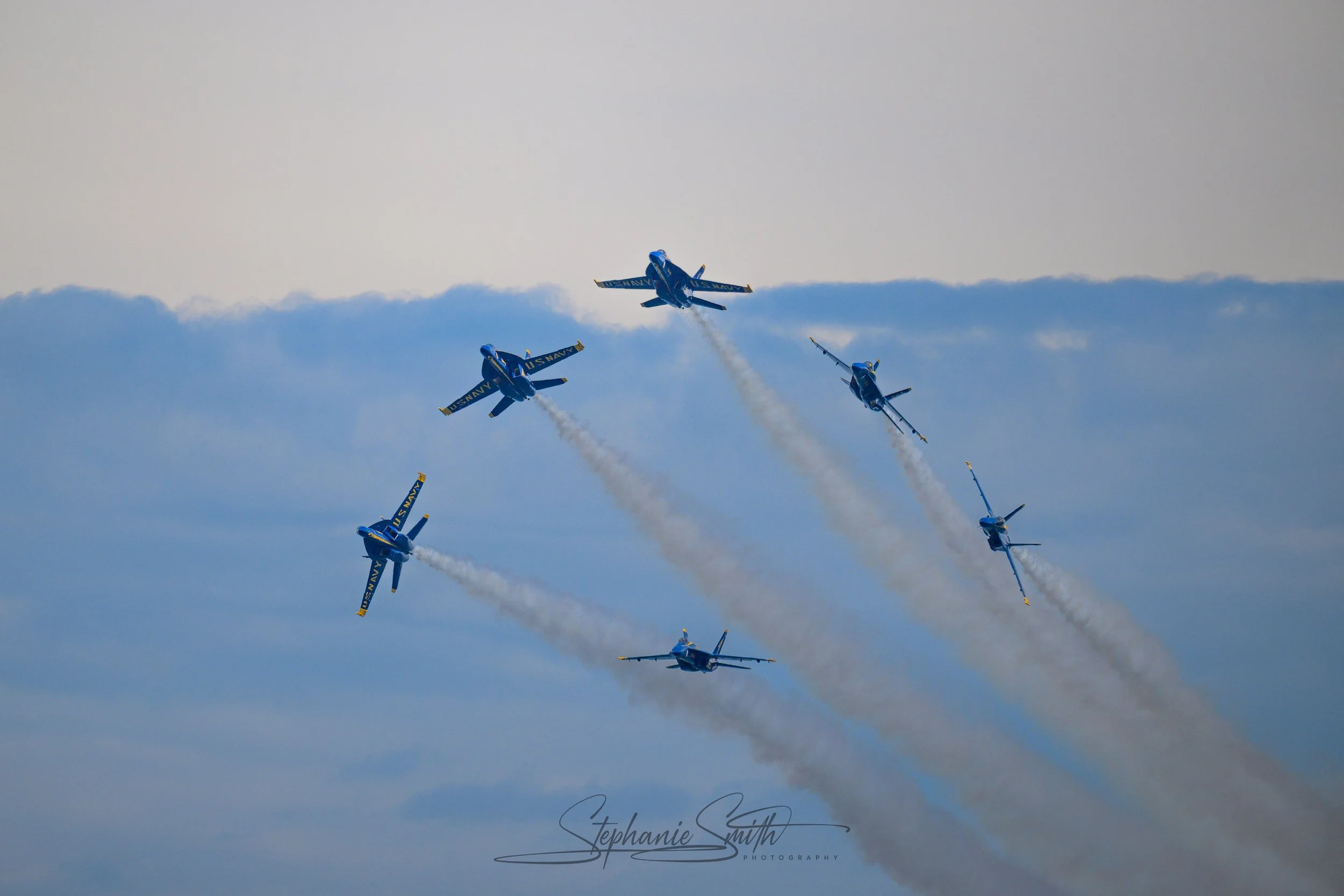 Blue U.S. Navy fighter jets flying in formation with smoke trails against a cloudy sky.