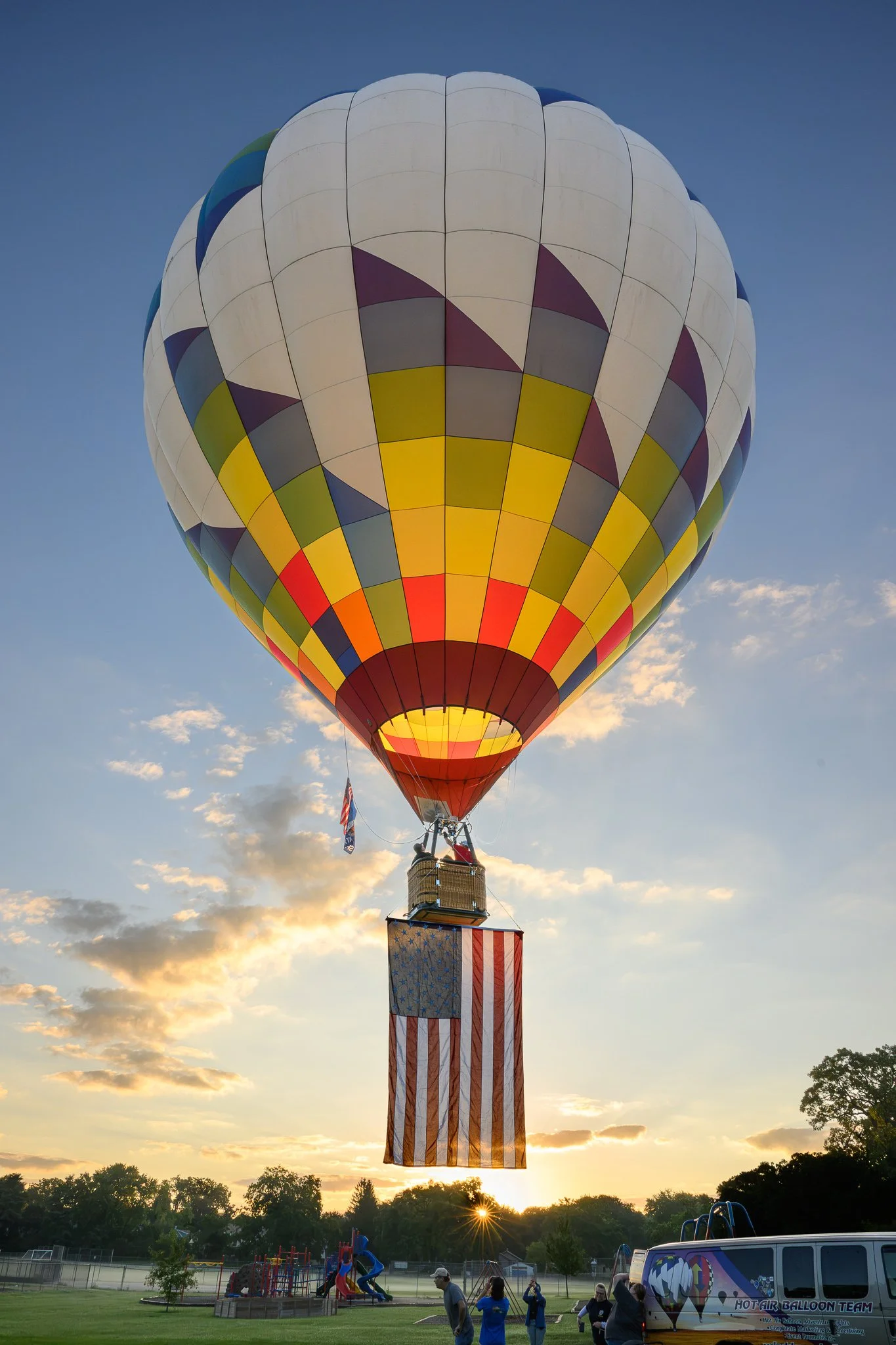 A colorful hot air balloon with a rainbow plaid pattern rising into the sky during sunset, with a group of people and a van on the ground nearby.