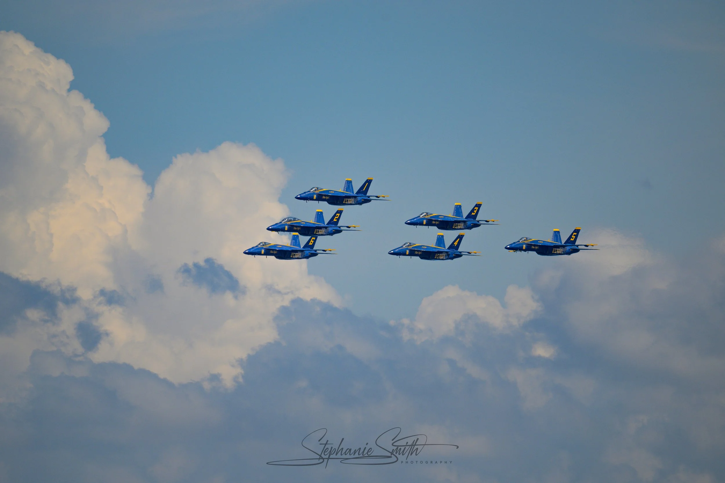 Six blue fighter jets flying in formation against a partly cloudy sky with white clouds.