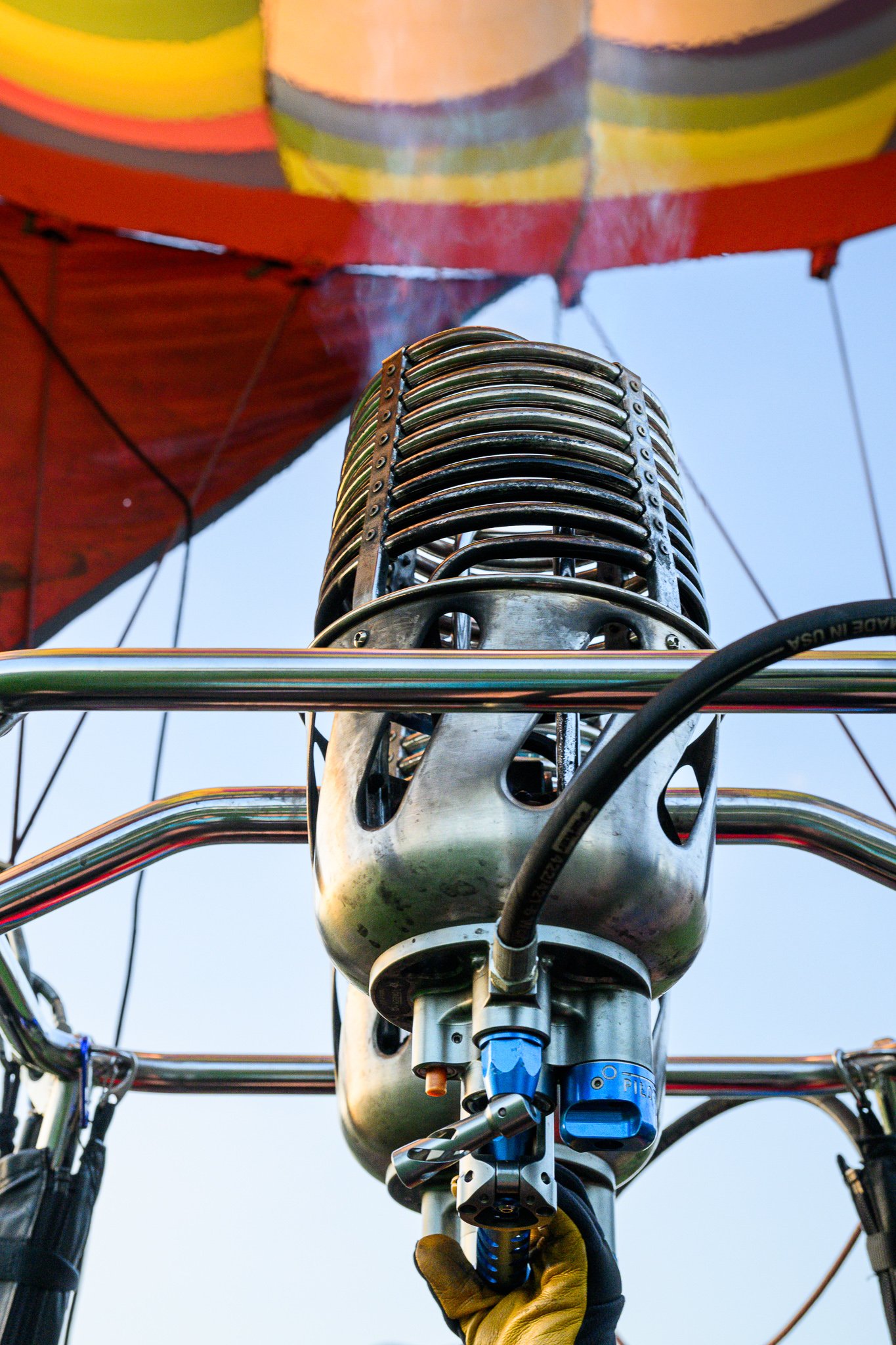Close-up view of a hot air balloon burner assembly, with a person wearing yellow gloves operating it while the balloon's colorful fabric is visible in the background.