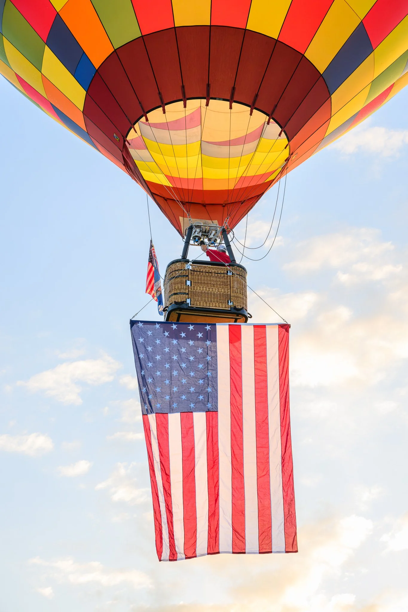 A hot air balloon with an American flag hanging from the basket, flying in a partly cloudy sky.