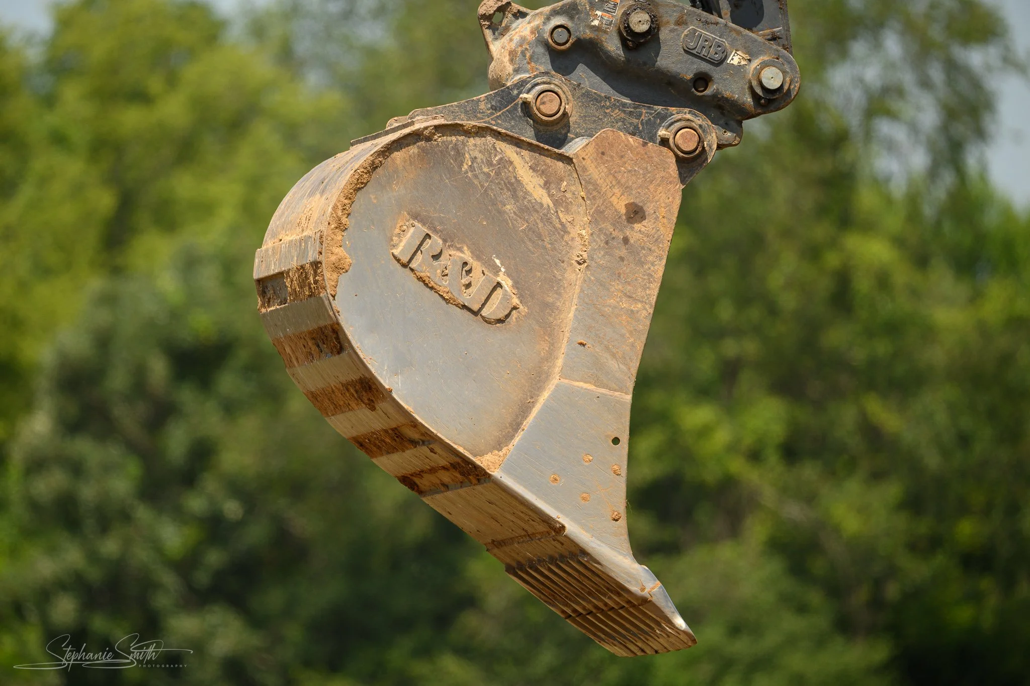 Close-up of a JRB hydraulic excavator bucket attached to an excavator arm, with a background of green trees, showing signs of use and dirt on the metal surface.