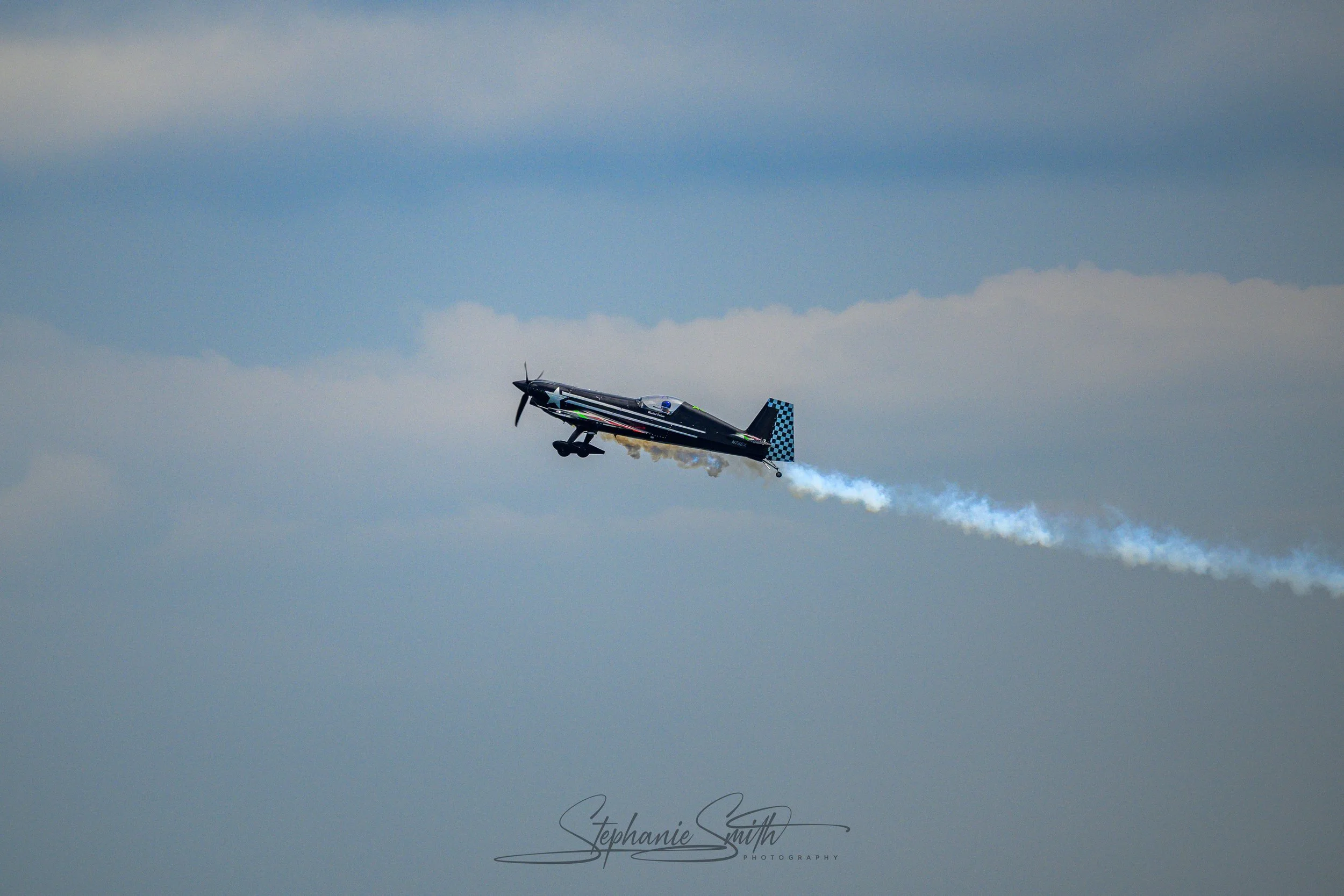A black military aircraft with blue checkered tail flying in a cloudy sky, releasing white smoke.