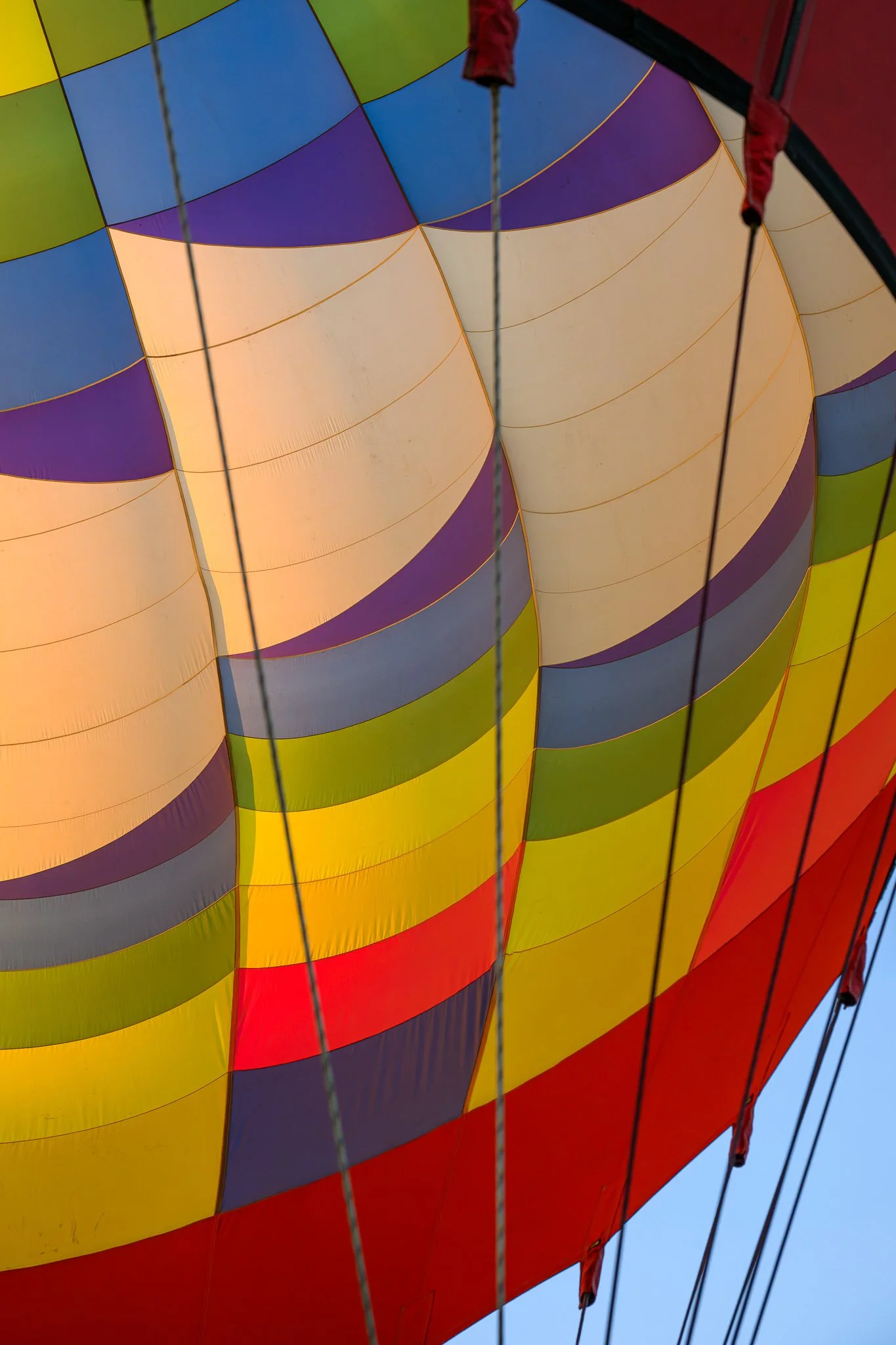 Close-up of a hot air balloon's colorful fabric panels, including yellow, blue, purple, and red sections, with supporting ropes and a clear blue sky in the background.