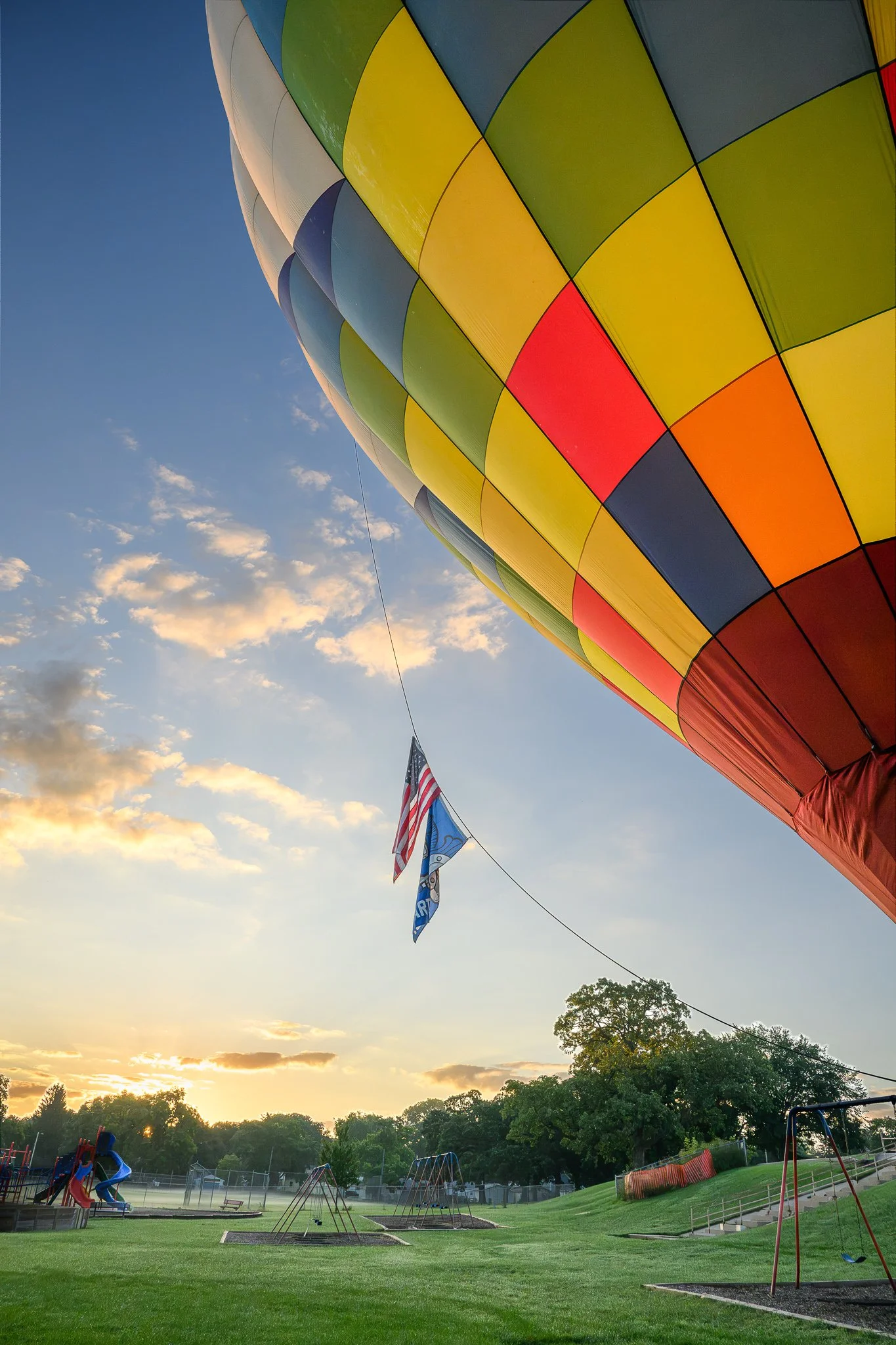 A colorful hot air balloon with a patchwork pattern, ballooning over a park at sunset with children playing on swings and a line of flags in the background.