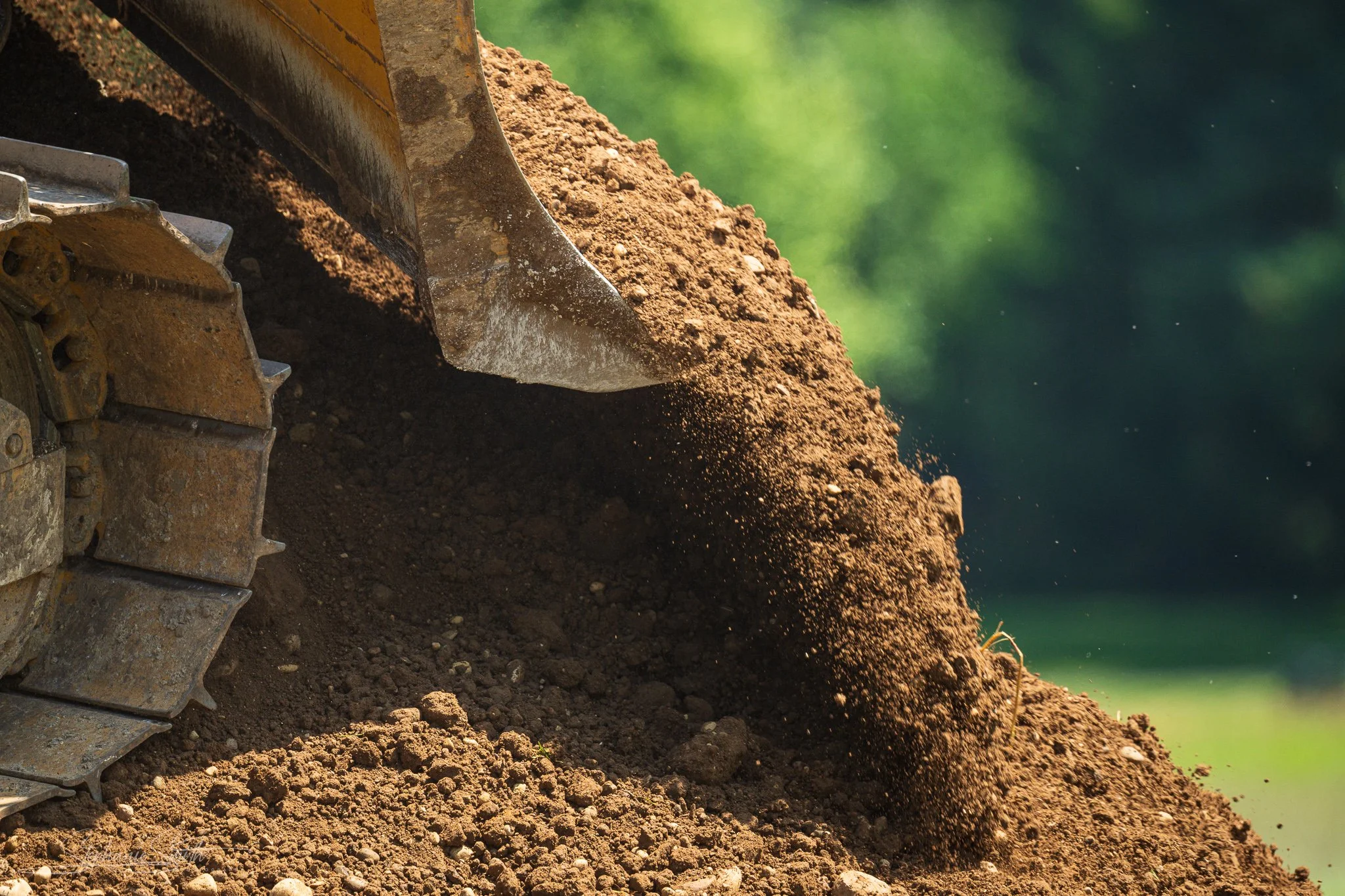 Close-up of a yellow bulldozer blade pushing a mound of dirt with a green blurred background.