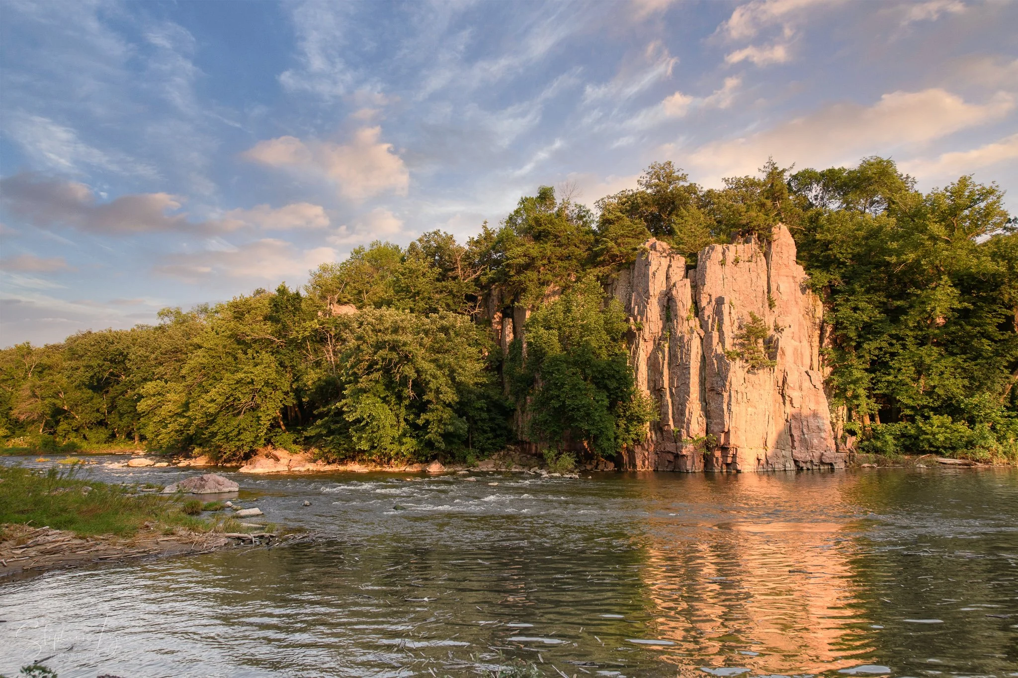 A river flowing at the base of a rocky cliff, with trees covering the top and a partly cloudy sky overhead.