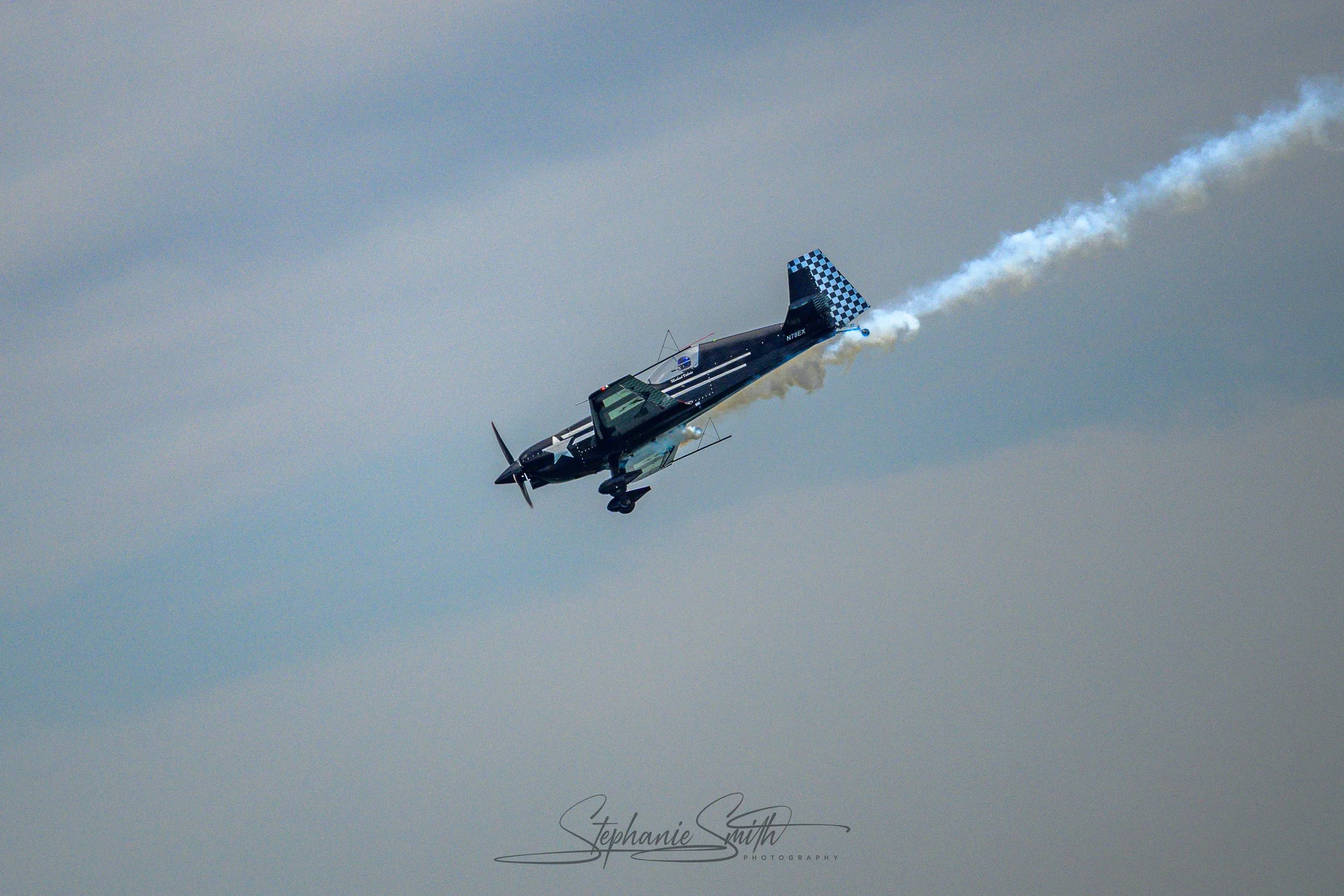 A blue and black fighter jet performing a dive, leaving white smoke trail behind against a cloudy sky.
