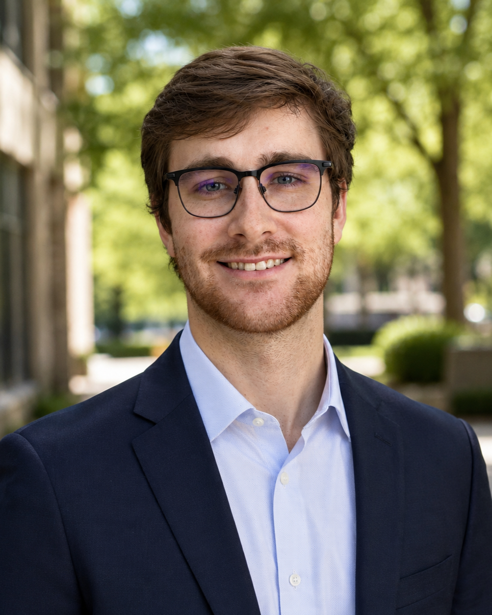 A young man with brown hair and glasses, smiling, wearing a navy blazer and light blue shirt, standing outdoors with blurred trees and buildings in the background.