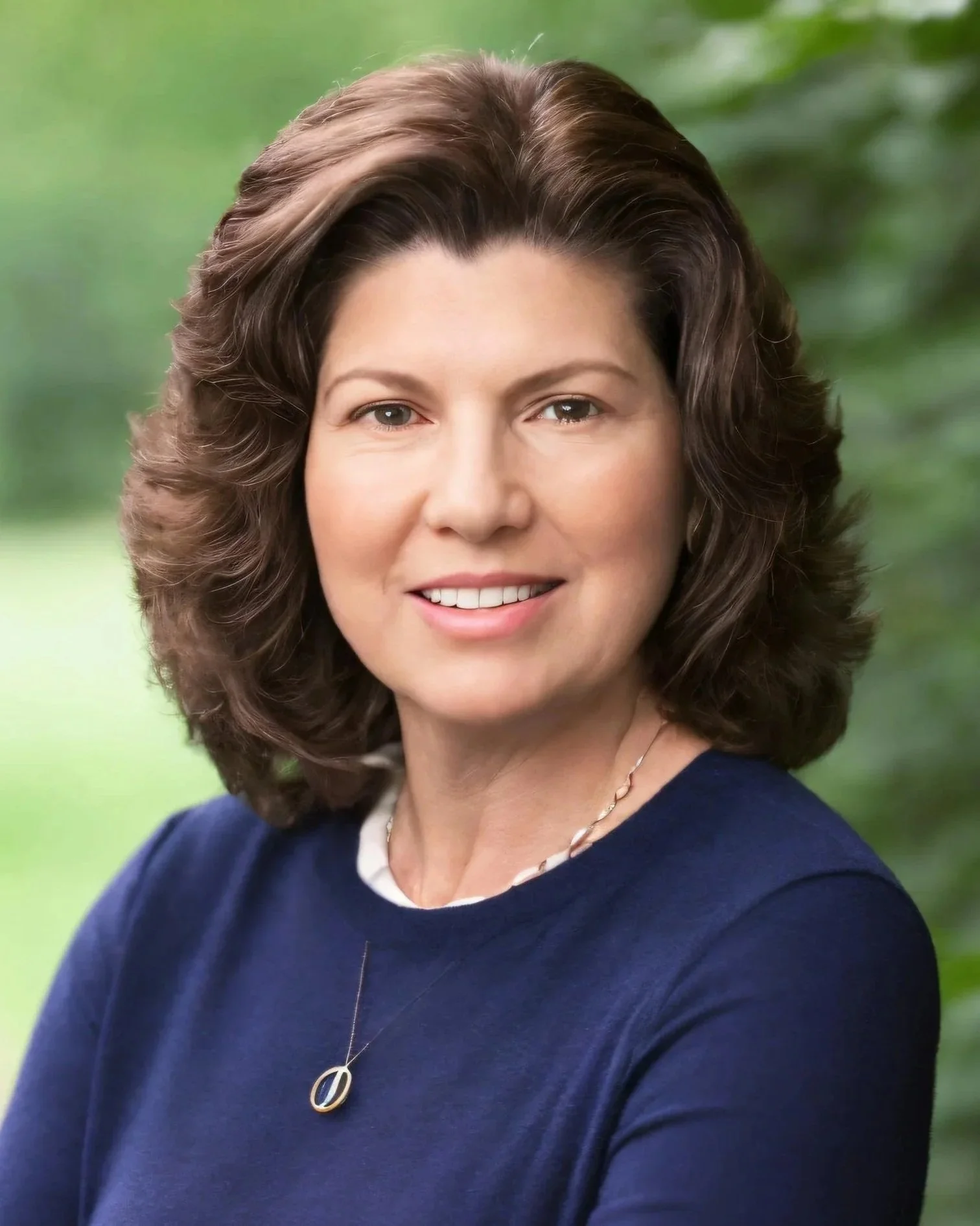A woman with shoulder-length brown hair, smiling, wearing a navy blue top, a necklace, and an oval pendant, standing outdoors with green blurred foliage in the background.