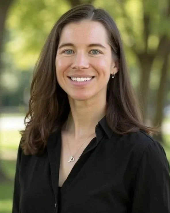 A woman outdoors with green trees in the background, wearing a black shirt and jewelry, smiling at the camera.