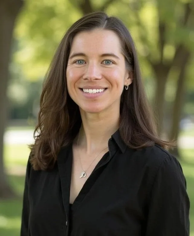 A woman with long brown hair and blue eyes smiling outdoors, wearing a black shirt and a silver necklace, with a background of green trees and sunlight.