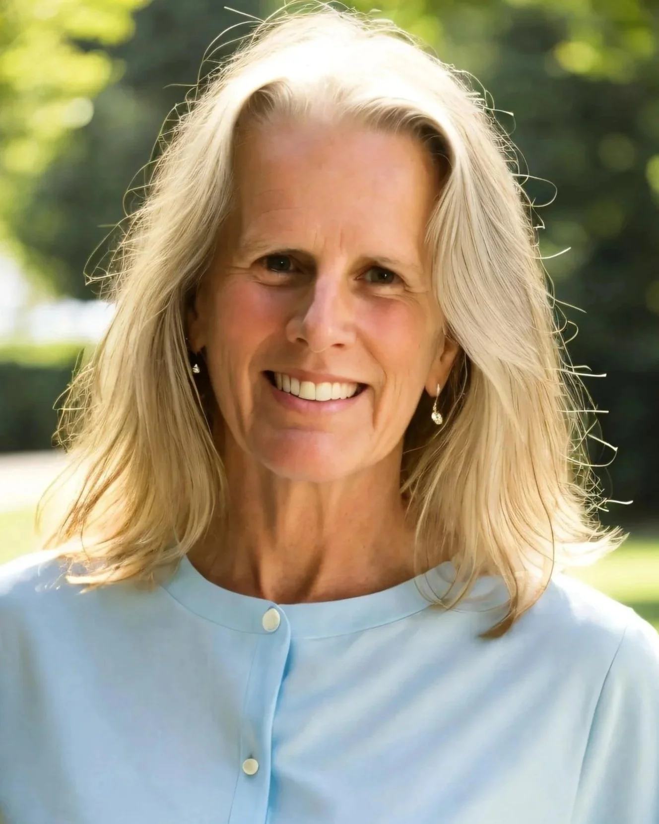 A woman with long blond hair and a warm smile, wearing a light blue blouse and earrings, standing outdoors with greenery in the background.