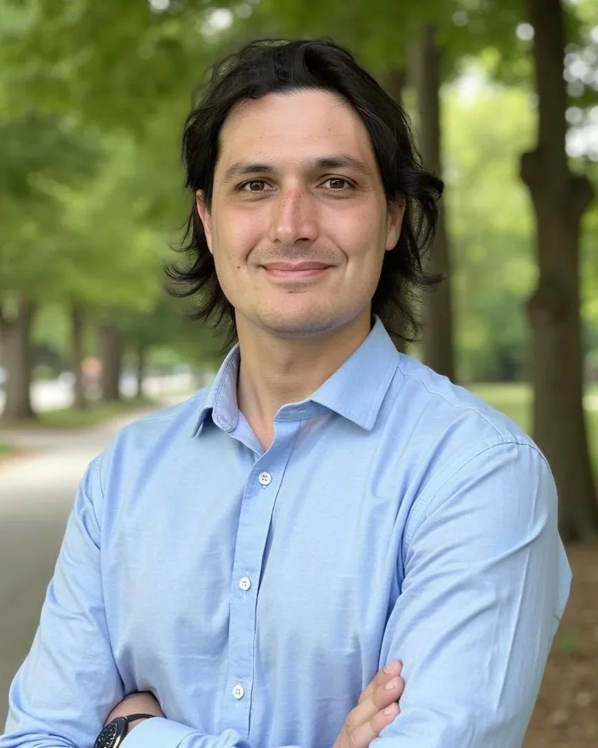 A young man with black, shoulder-length hair, wearing a light blue button-up shirt, stands outdoors in a park with green trees in the background. He is smiling slightly and has his arms crossed.
