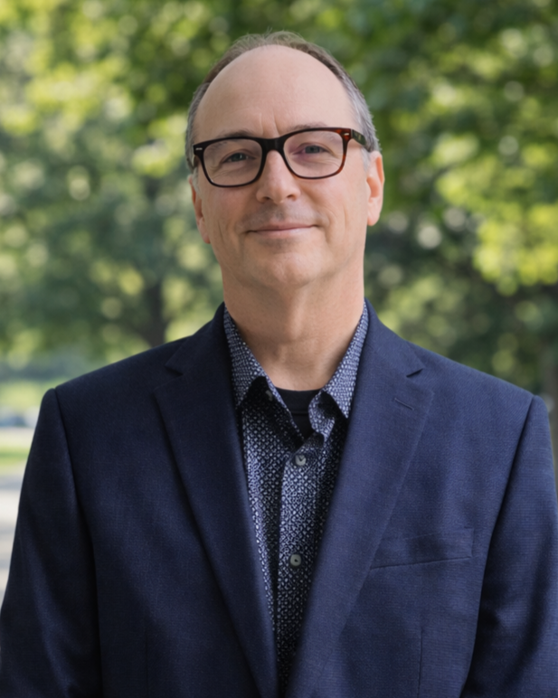 A middle-aged man in a navy blazer, patterned shirt, and glasses standing outdoors with trees and greenery in the background.