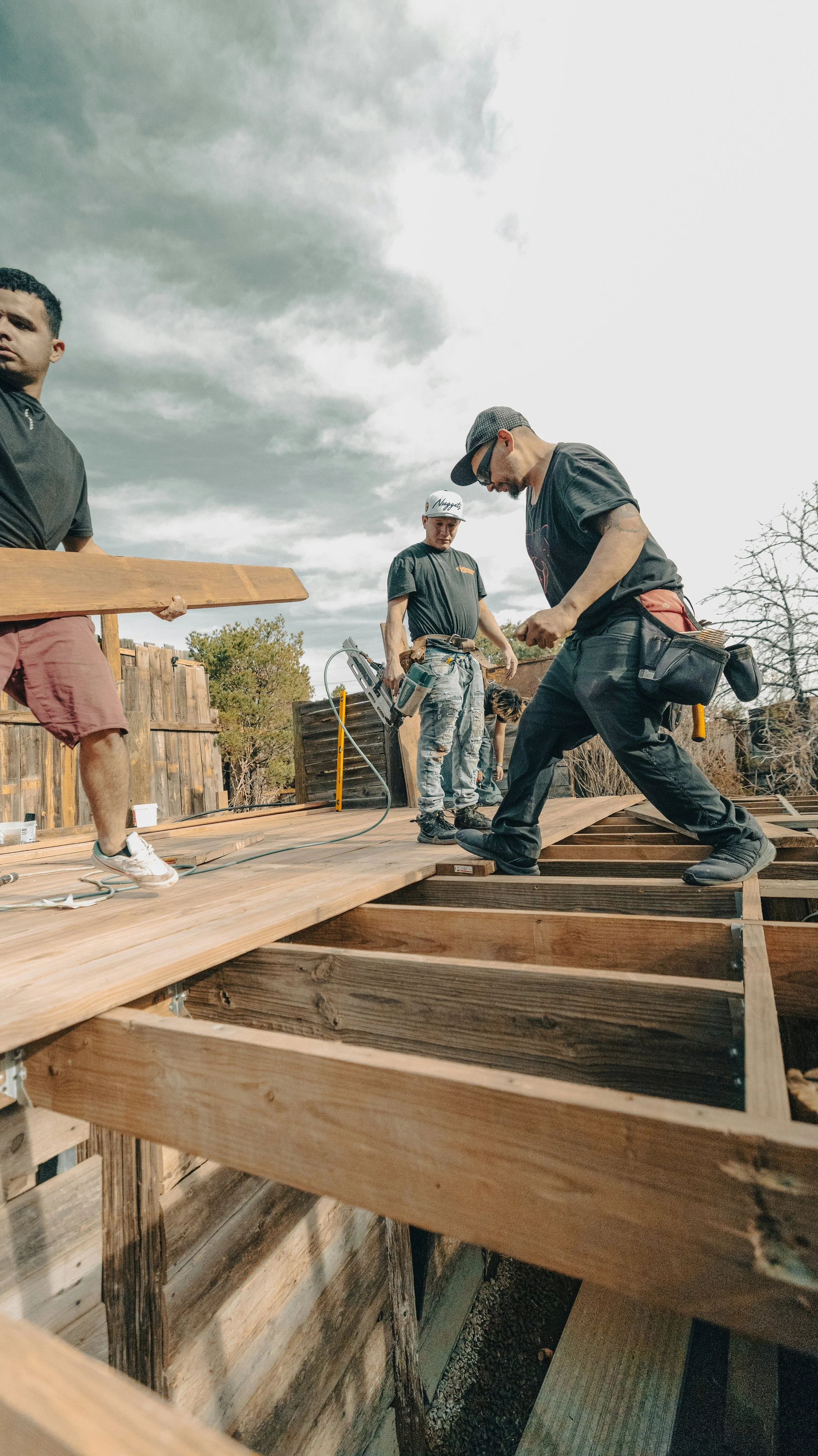 Three men are working on a wooden deck construction outside, with cloudy skies overhead.