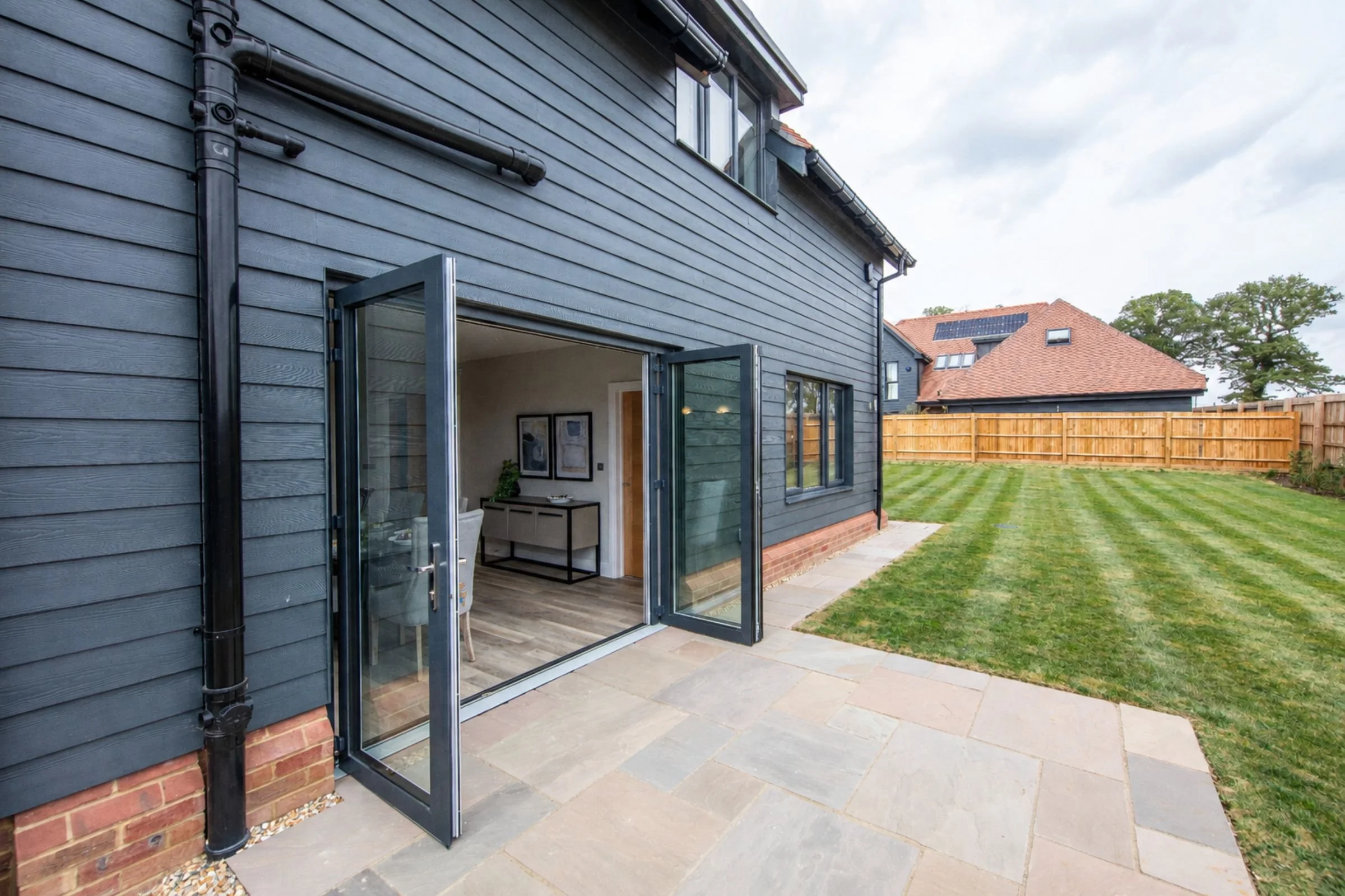 Paved rear terrace and lawn beside open bifold doors to kitchen/diner.