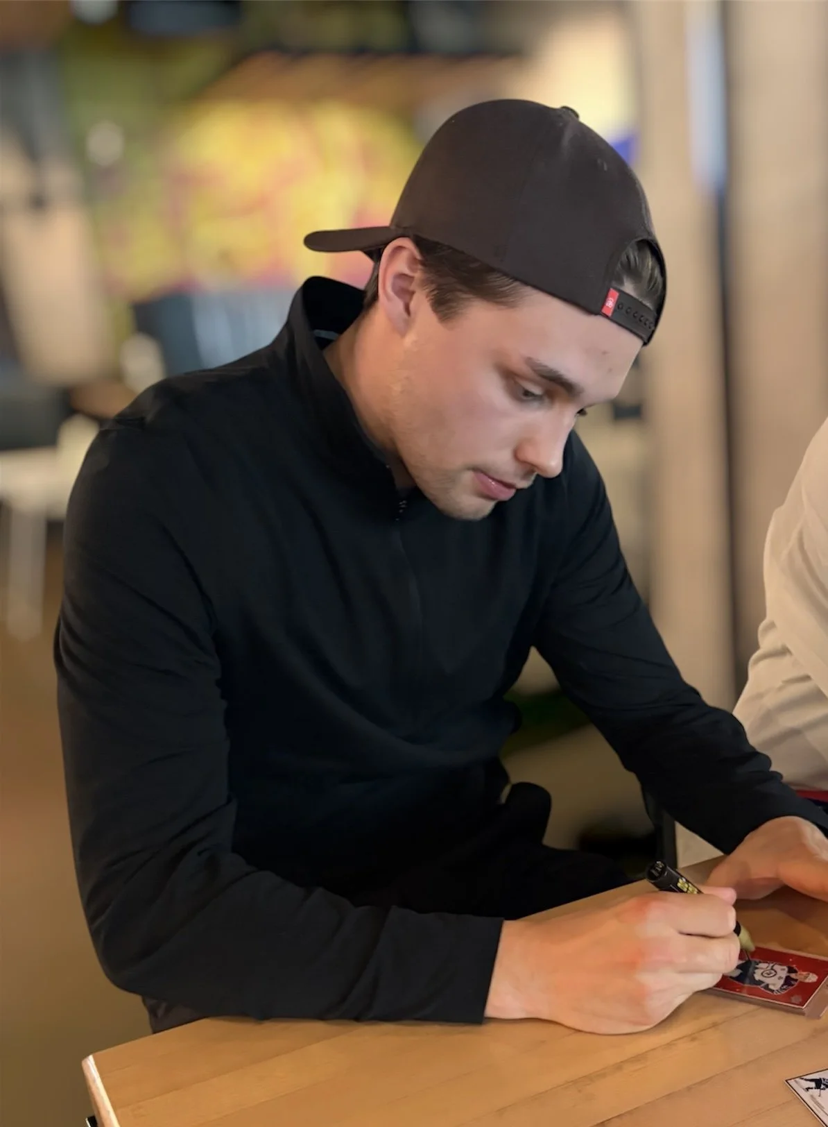A young man in black clothing and a backwards black baseball cap, sitting at a wooden table, is signing an autograph with a black marker. The background is blurred, showing a colorful, out-of-focus decor.