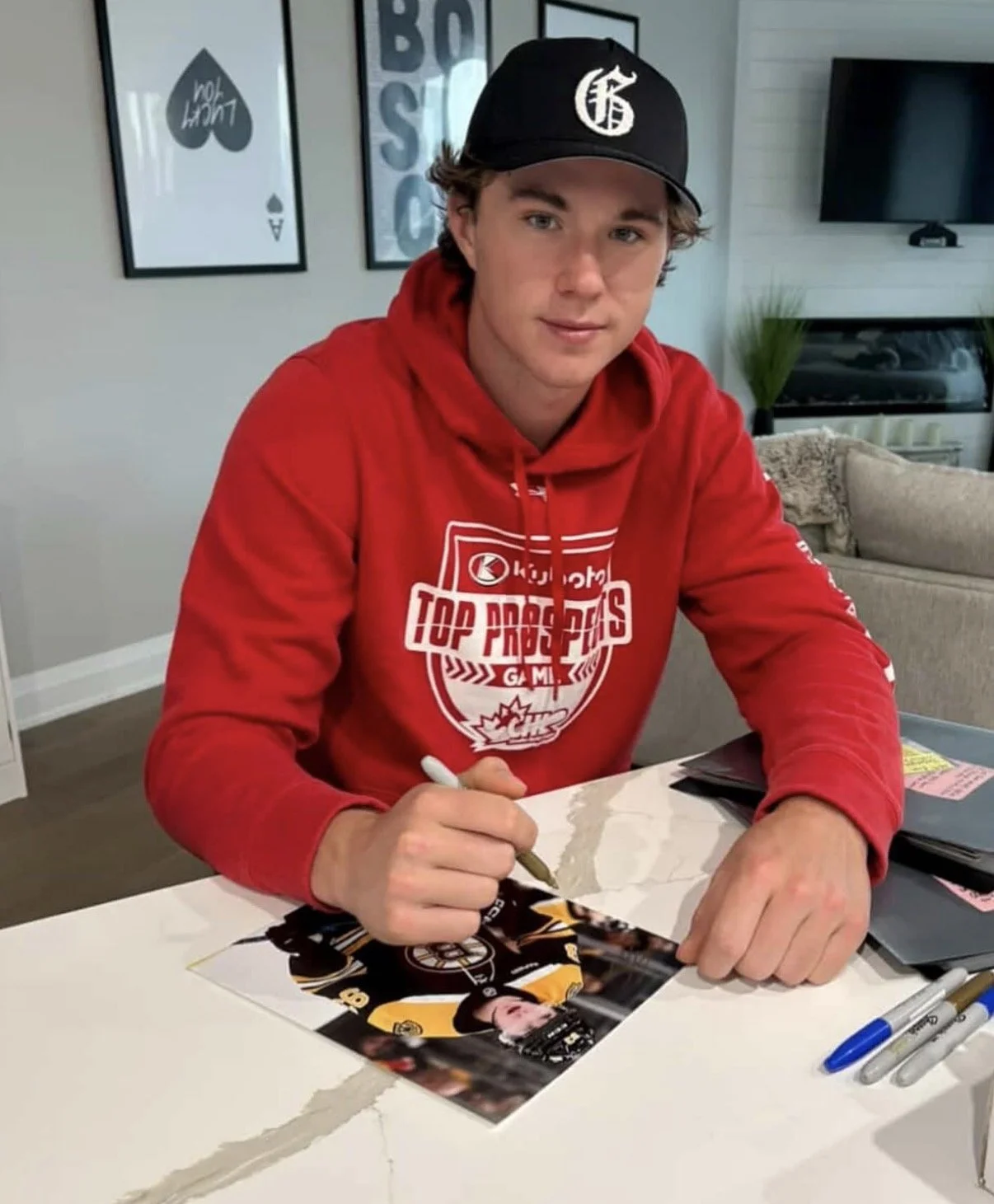 A young man with light skin and curly brown hair, wearing a black cap with a white logo and a red hoodie with sports-themed text, is sitting at a white table signing a photograph of himself in a hockey jersey with a white marker. The background includes a wall with framed art prints, a flat-screen TV, and a sofa with a throw blanket.