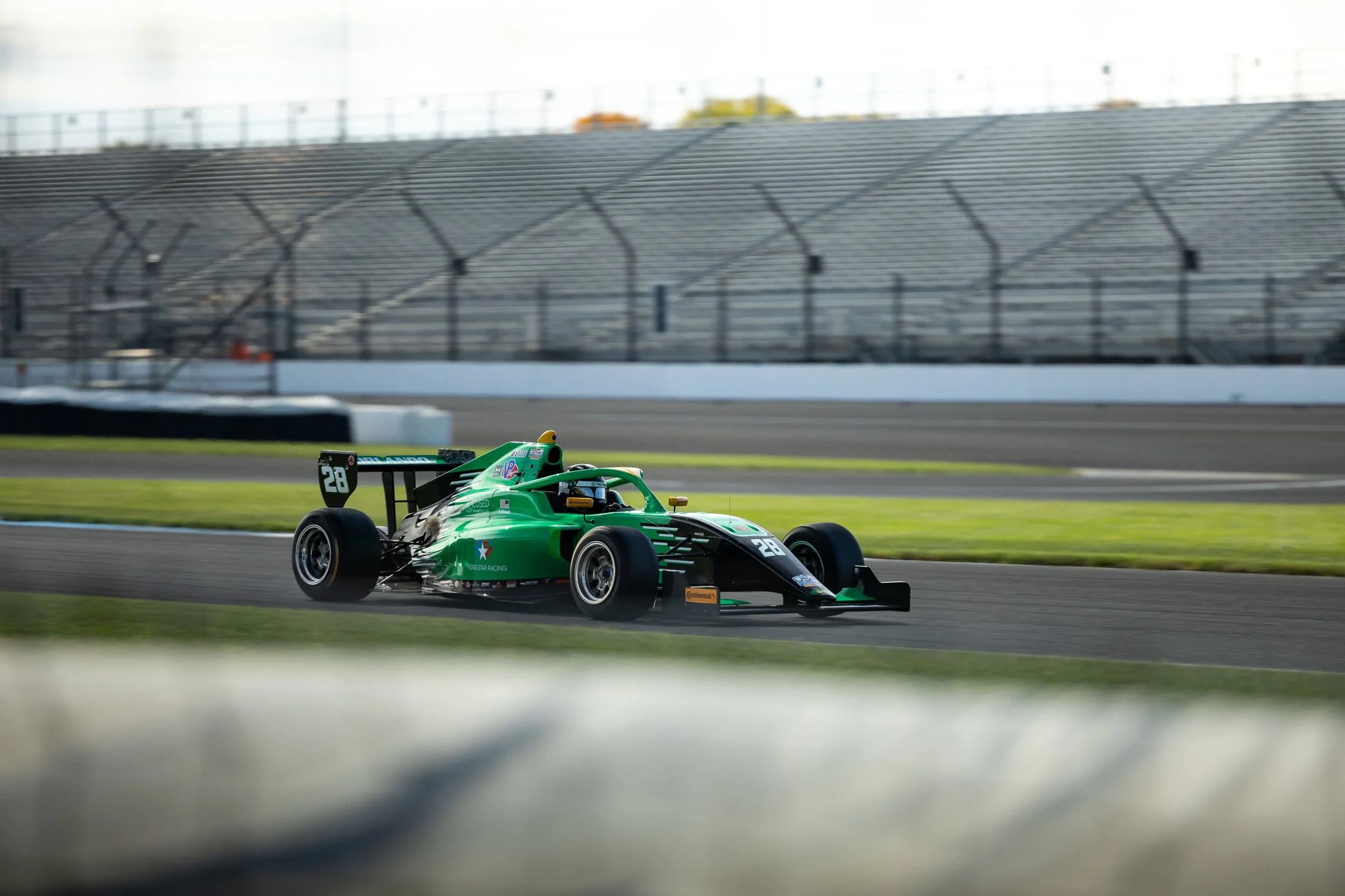 Nick d'Orlando green and black open-wheel race car driving on a race track, with grandstands in the background.