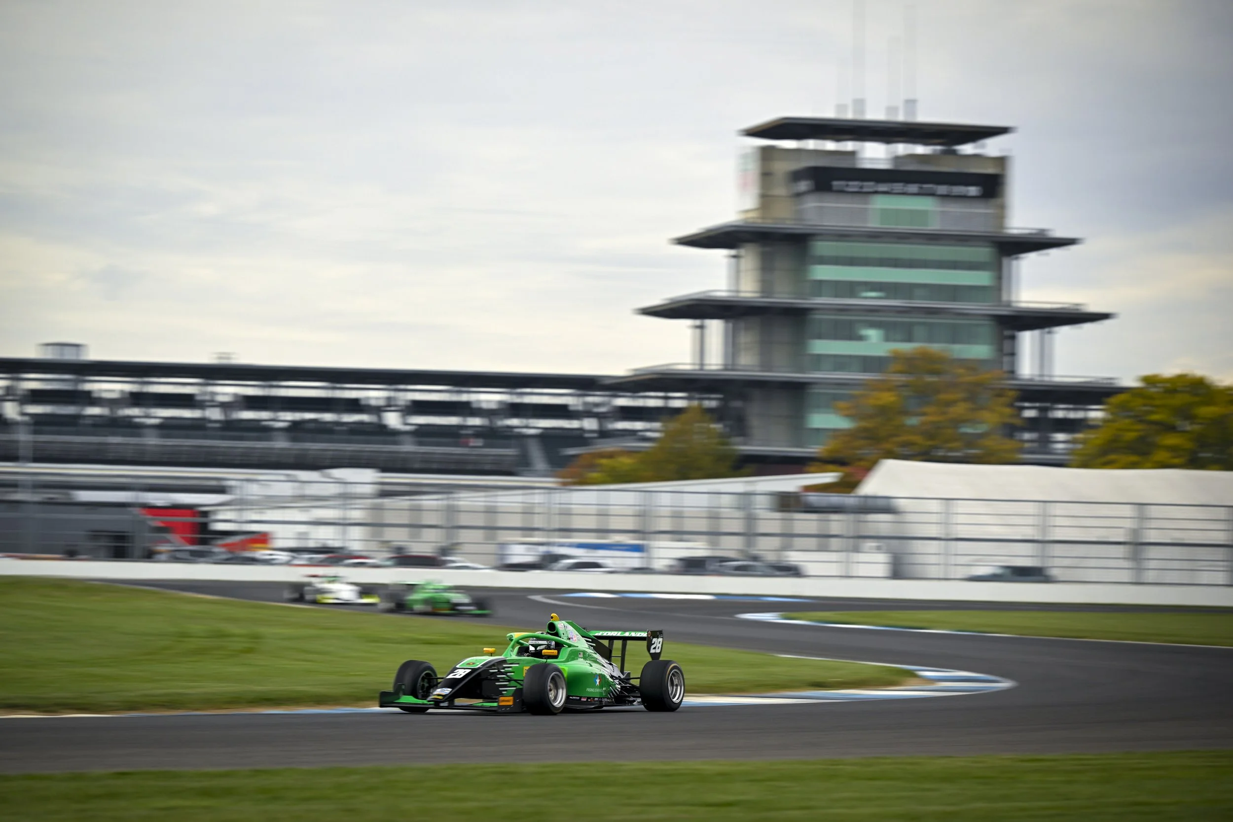 Nick d'Orlando in a green race car speeding on a race track with a blurred background of a large modern building and other race cars in the distance.