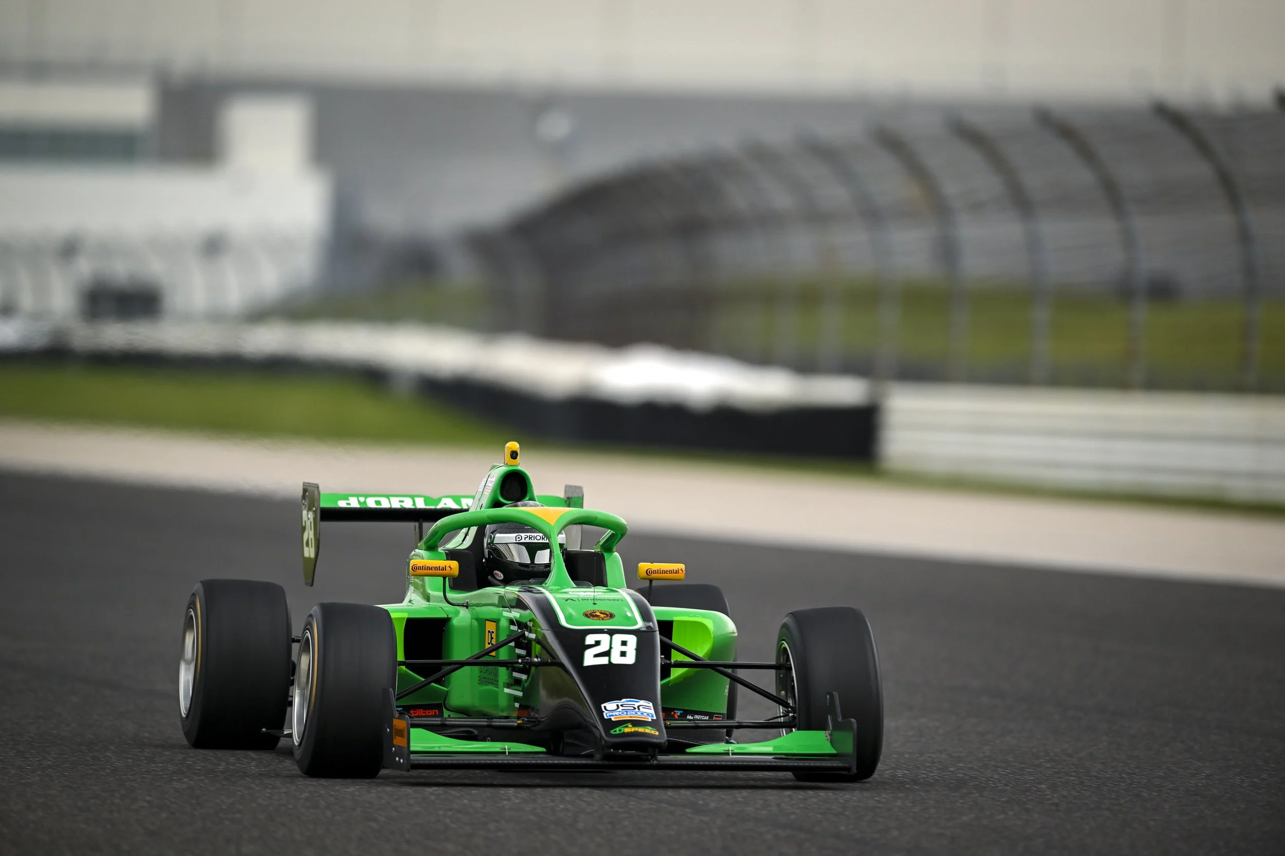 Nick d'Orlando green open-wheel race car on a racetrack, with a blurred grandstand and track in the background.