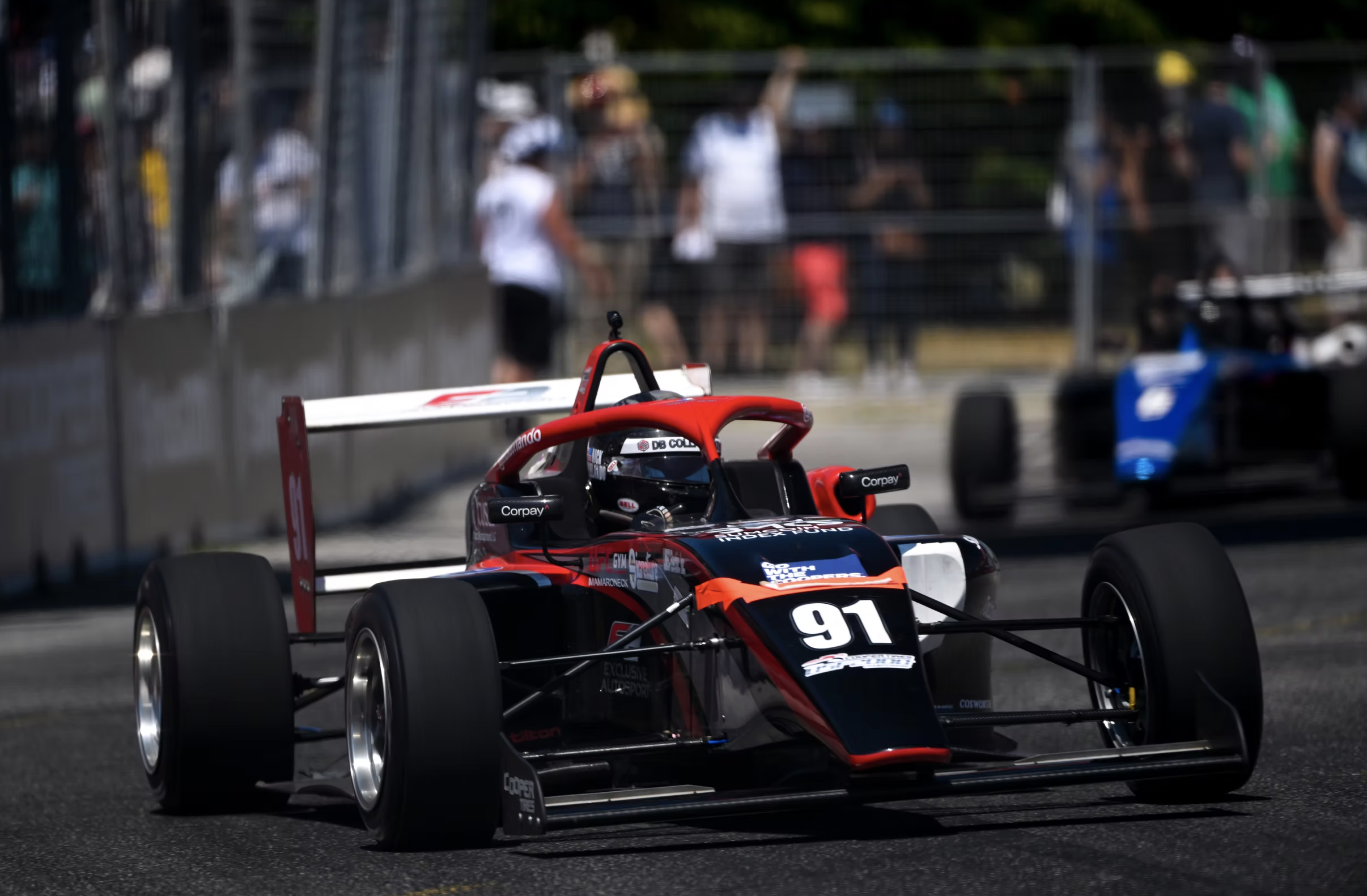 Nick d'Orlando in a red and black Formula racing car on a race track with a blurred background of spectators and team members behind a fence.