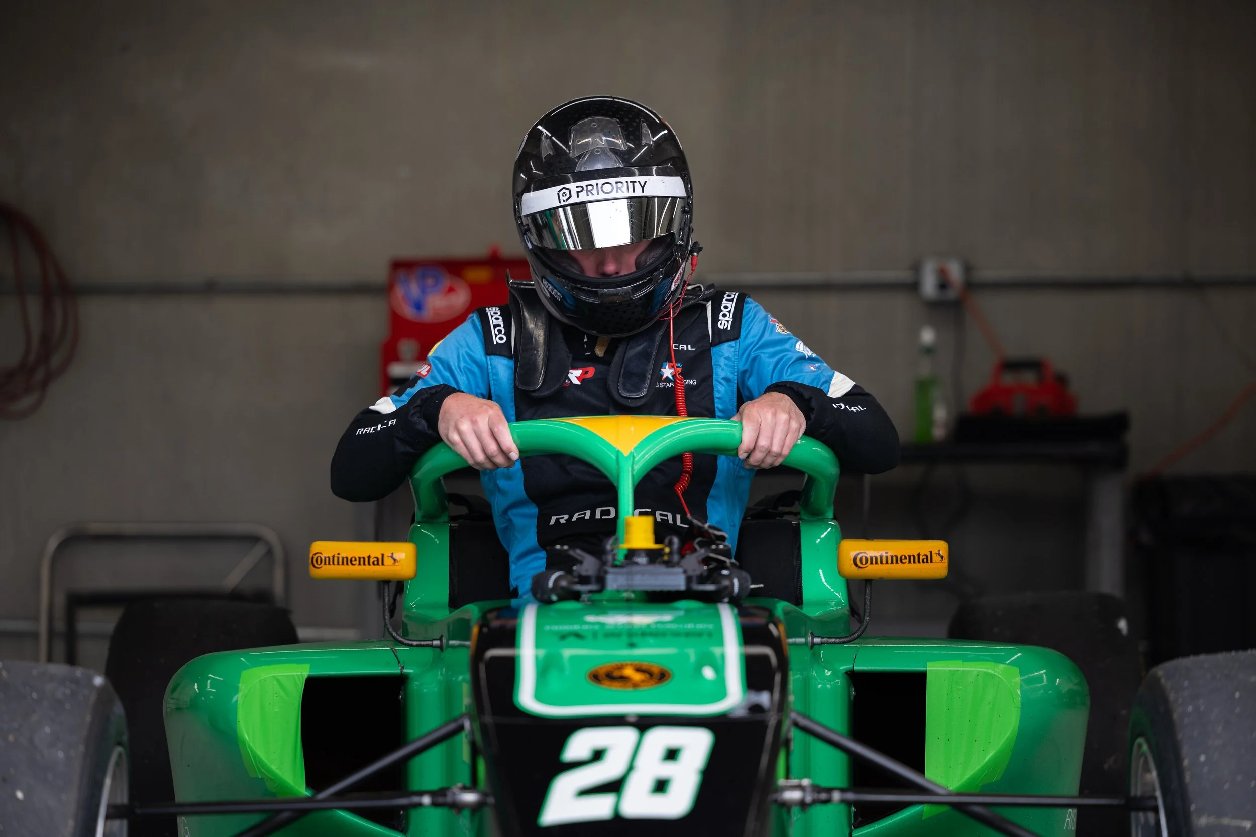 Nick d'Orlando professional race car driver in a helmet and racing suit preparing in a garage, sitting behind the steering wheel of a green race car with the number 28.