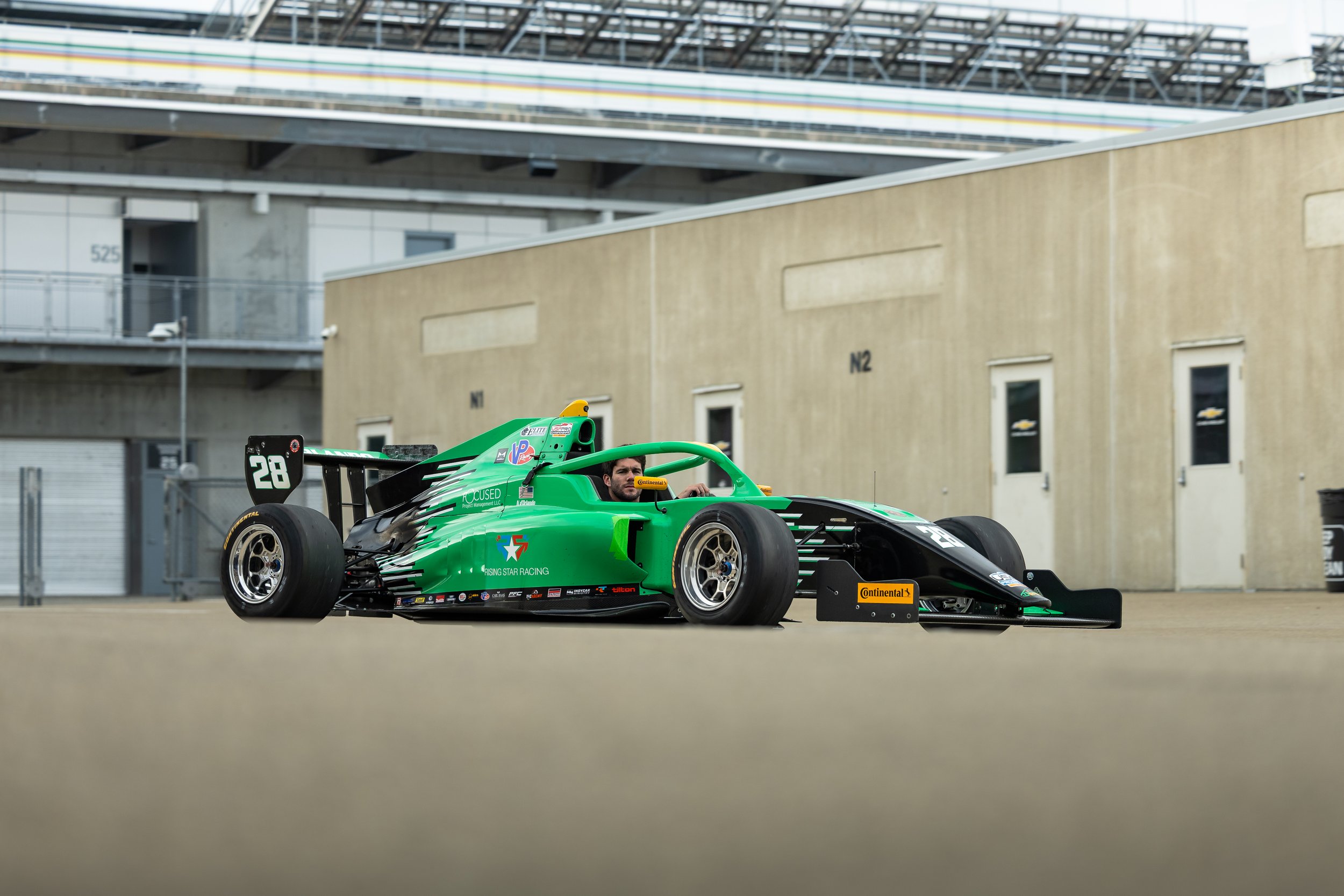 Green Formula race car on a racetrack with driver in helmet, the background shows industrial buildings and overcast sky.