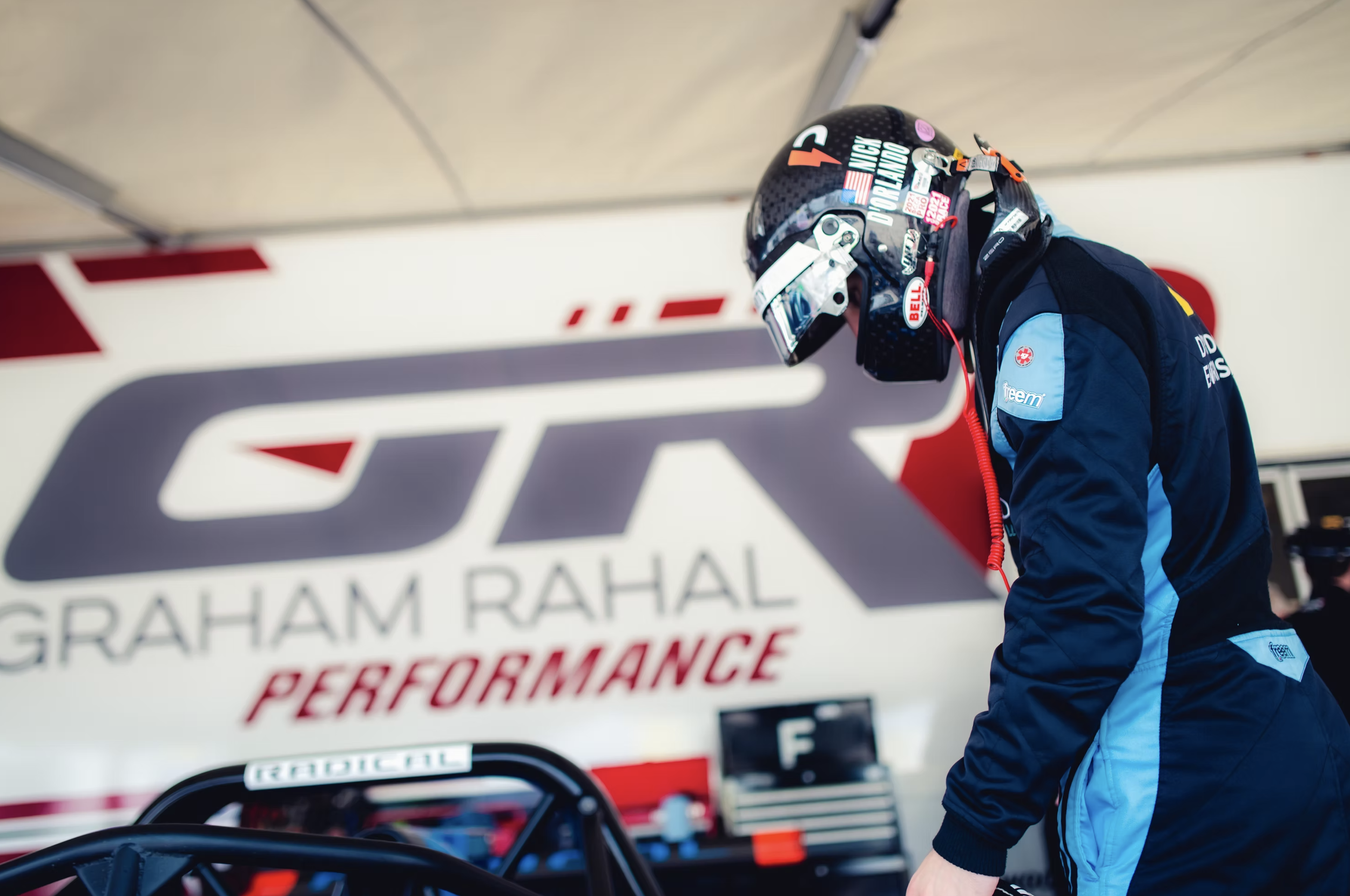 Nick d'Orlando in a black helmet with various stickers and a blue racing suit standing next to a race car and a large banner in the background with the words 'GRAHAM RAHAL PERFORMANCE'.