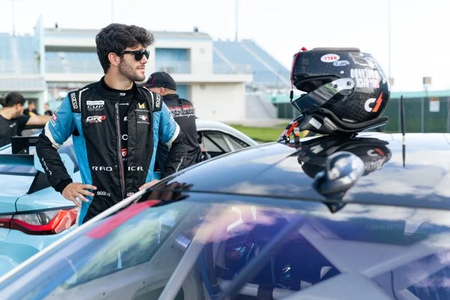 A race car driver wearing a racing suit and sunglasses stands next to a black race car with a helmet placed on the roof, at a racetrack.