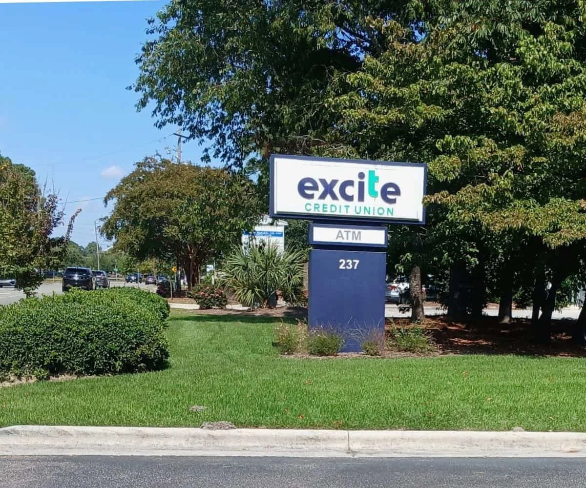 Sign for Excite Credit Union with ATM at 237, on a grassy area with trees and bushes, and a parking lot with cars in the background.