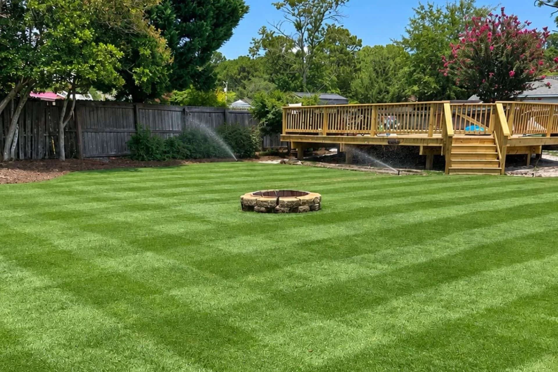 A backyard with a neatly mowed green lawn, a tree ring fire pit made of bricks, a raised wooden deck with stairs, a sprinkler spraying water, potted plants on the deck, a wooden fence, and trees with flowering bushes in the background.