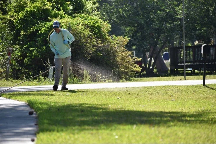 Turfology technician applying lawn treatment near a sidewalk on a tree-lined residential street in Wilmington, NC