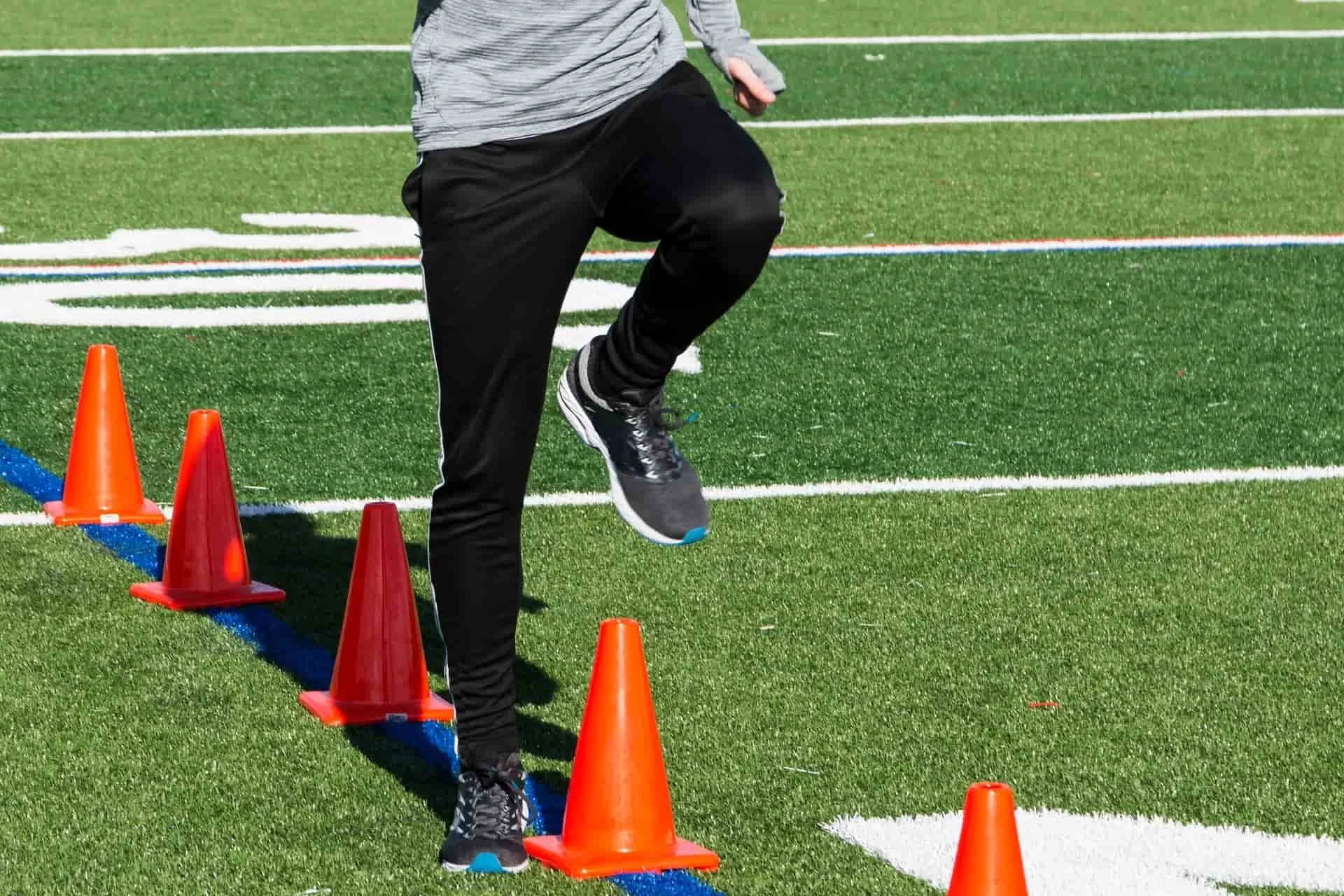 A person dressed in athletic clothing running through orange cones on a green turf field with white and blue painted lines.
