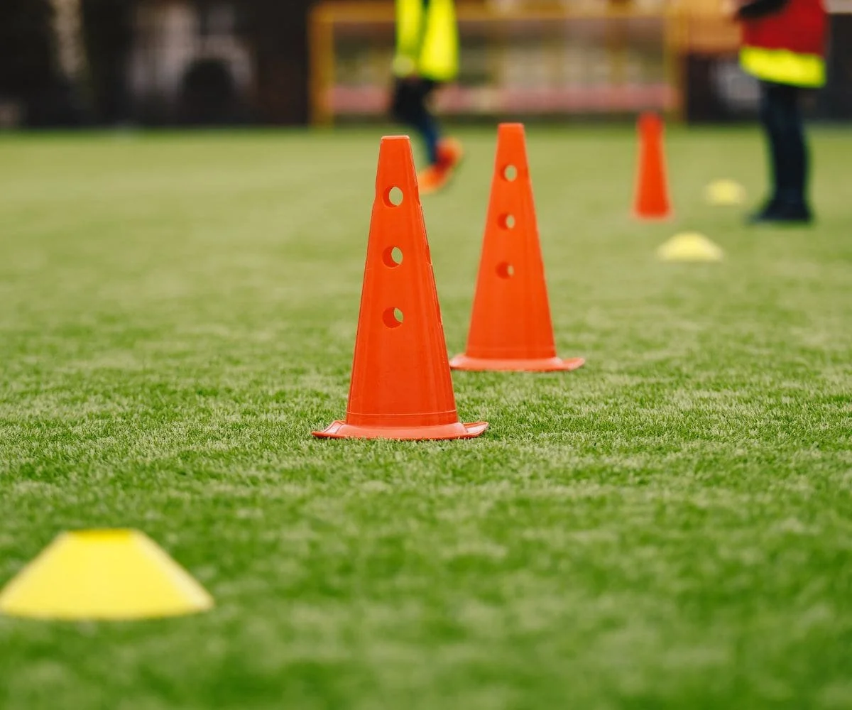 Orange traffic cones arranged on a grassy sports turf, used for sports drills or training, with blurred figures of trainers or players in the background.